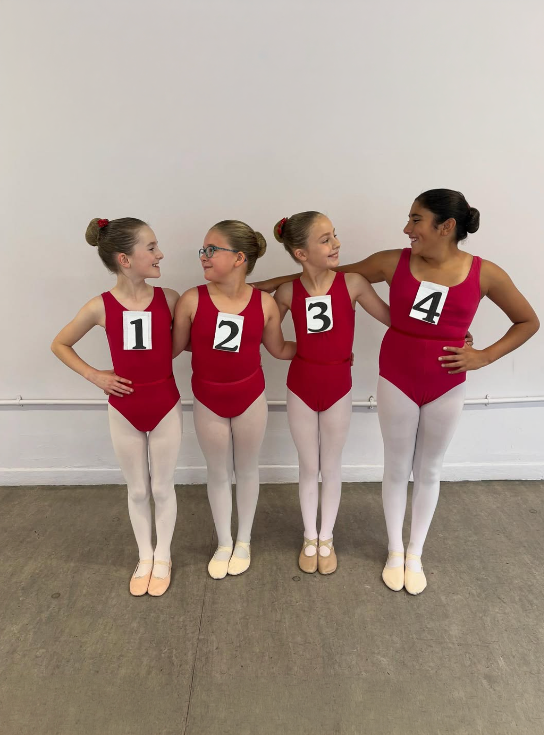 Four young girls dressed in red ballet leotards and white tights, standing side by side with their arms around each other, smiling. Each girl has a number pinned to their leotard, numbered 1 through 4. They are in a dance studio with a plain white wall and a ballet barre behind them.