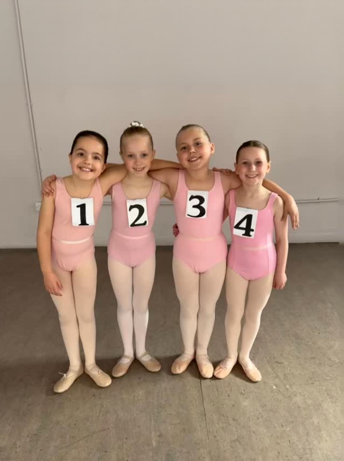 Four young girls in pink ballet costumes and tights standing together with arms around each other, smiling, each wearing a number on their chest from 1 to 4.