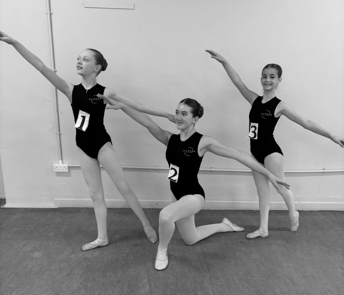 Three young female ballet dancers in black leotards, wearing numbers 1, 2, and 3, pose in a ballet studio with a plain wall and electrical outlets in the background, practicing ballet poses.