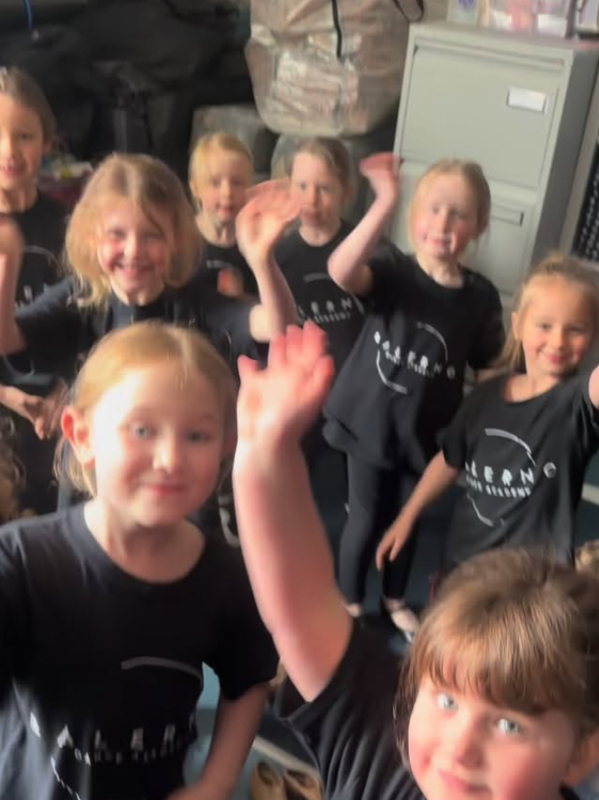 A group of young children, mostly girls, wearing black T-shirts with a white logo, smiling and waving at the camera inside a room that contains a filing cabinet and backpacks.