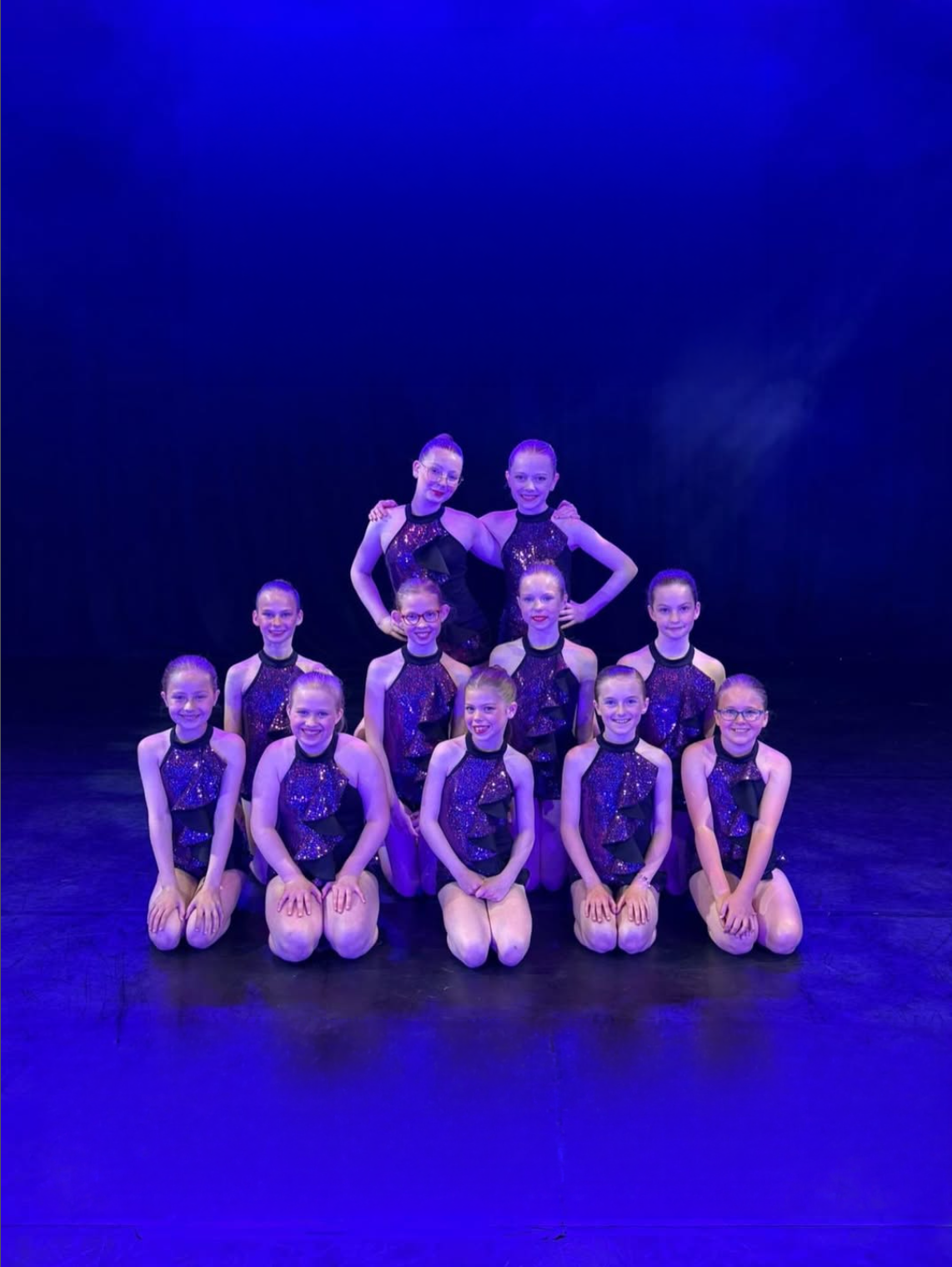Group of ten young girls in purple and black dance costumes, posing on stage under purple lighting.