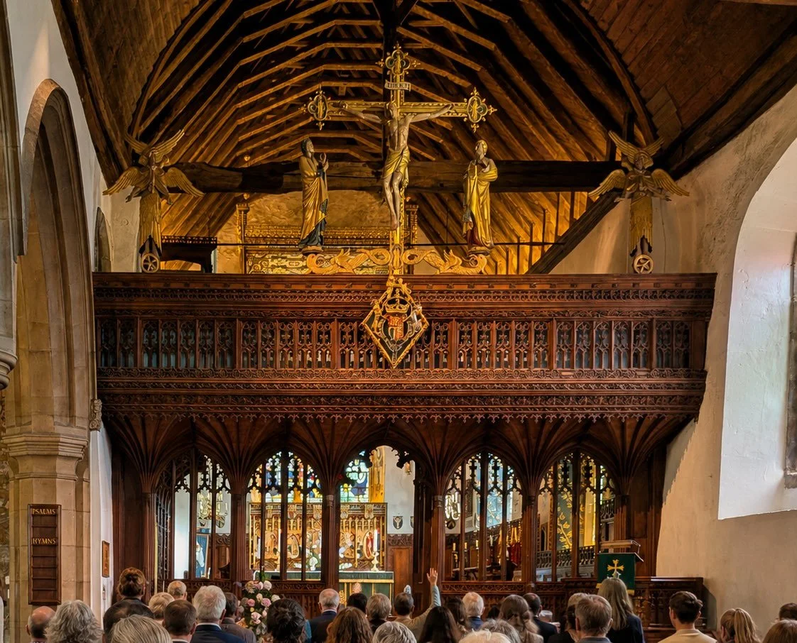 Interior of a church with a wooden altar and stained glass windows, congregation seated, some raising their hands, crucifix with Jesus and statues of angels or saints above.