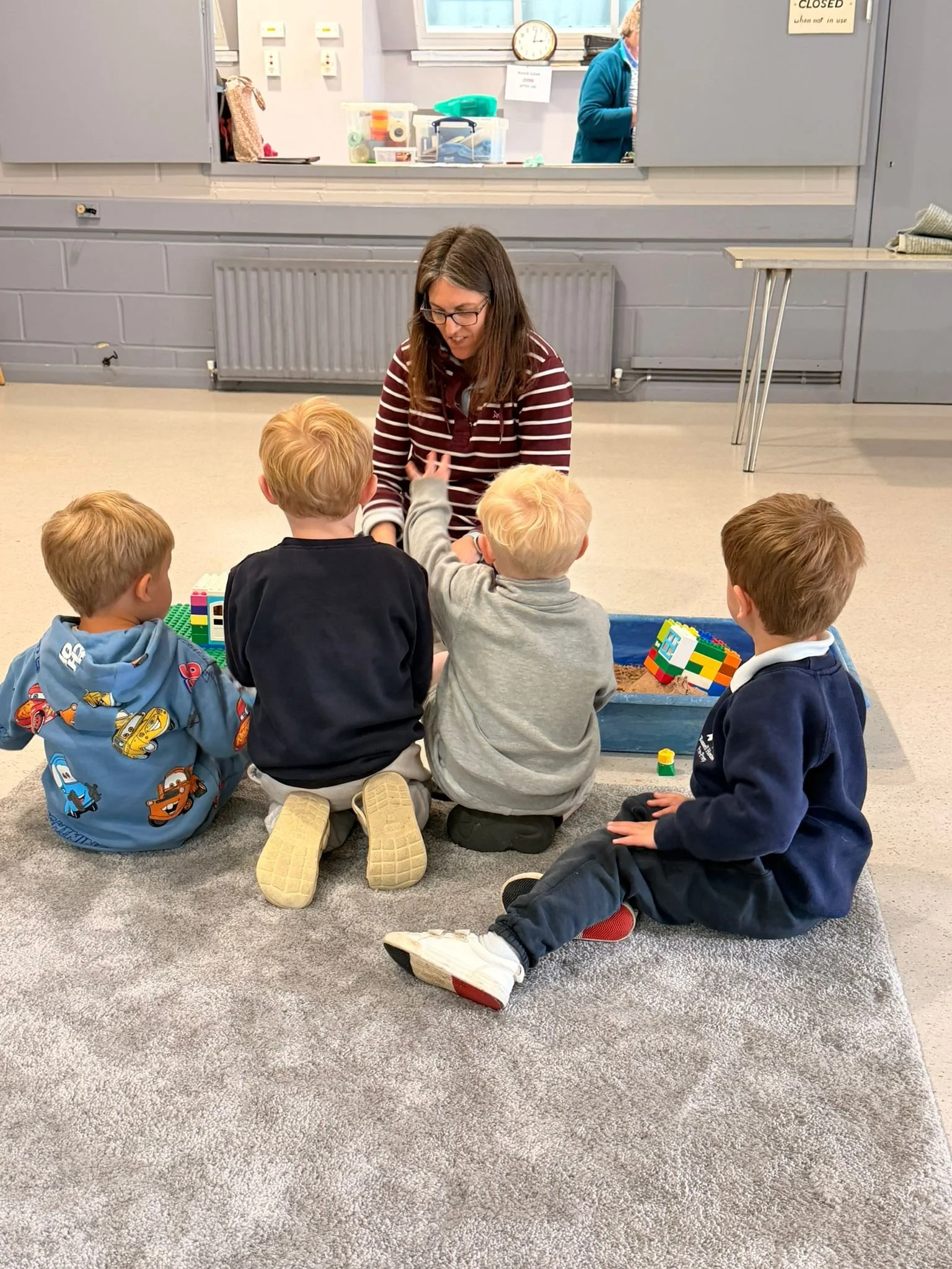 A woman is sitting on the floor with four young boys. The boys are playing with LEGO bricks, and the woman appears to be interacting with them. There is a window and a counter in the background, with a person visible behind the window.