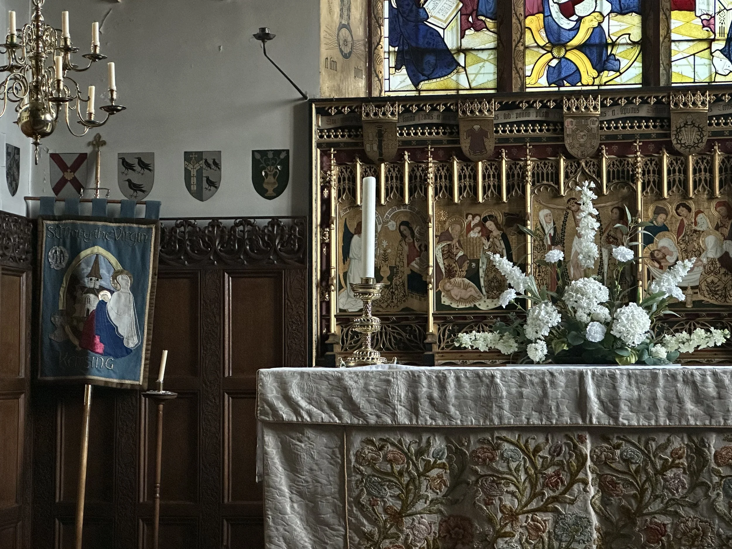 The interior of a church with stained glass windows depicting religious figures and scenes, shields with heraldic symbols on the wall, and an altar decorated with white flowers.