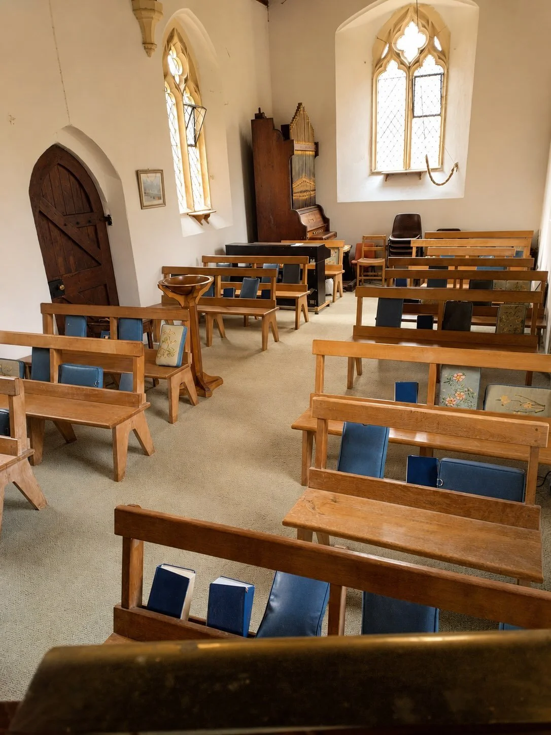 Interior of a small church or chapel with wooden pews, a pipe organ in the corner, stained glass windows, and a baptismal font.