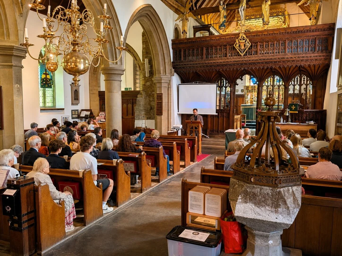 A man giving a speech or presentation at a church or cathedral with a large audience seated on wooden pews, classic chandeliers, stained glass windows, and an ornate wooden altar in the background.