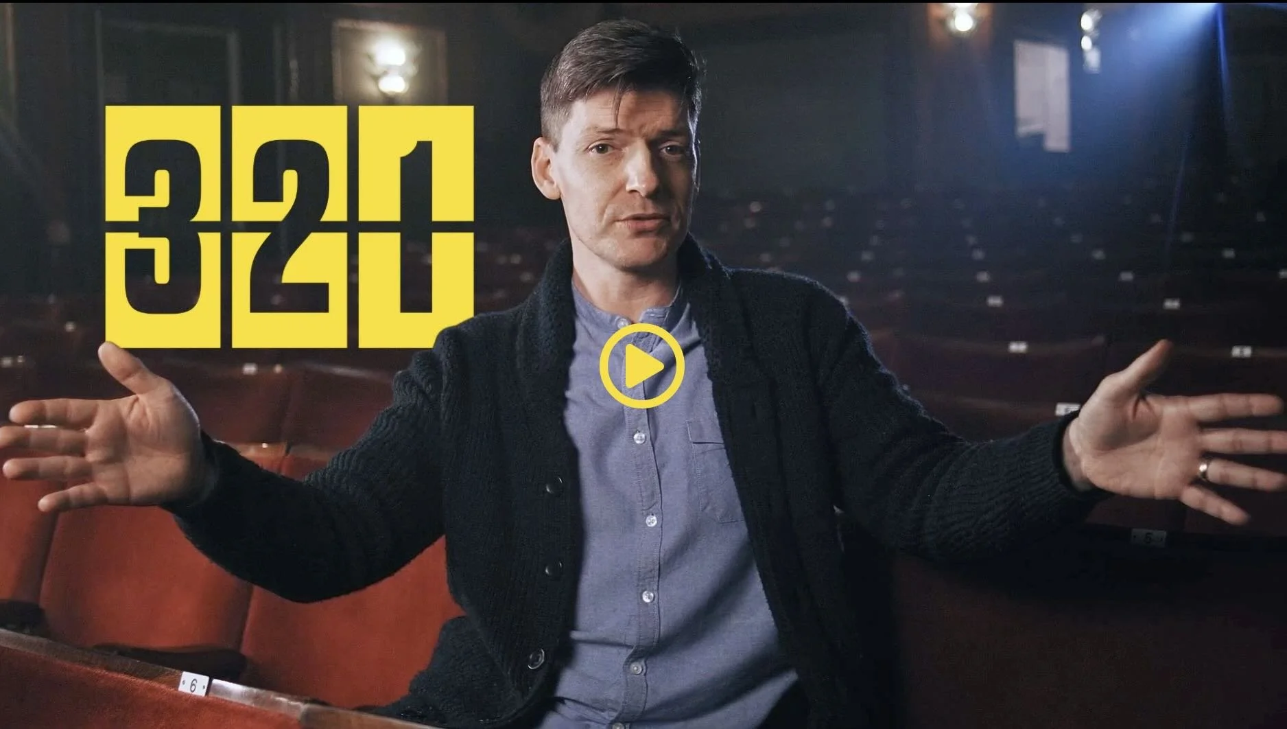 A man sitting in an empty theater with red seats and a dark background, gesturing with his hands, wearing a black sweater and a gray shirt, with large yellow text and a play button overlay on the image.