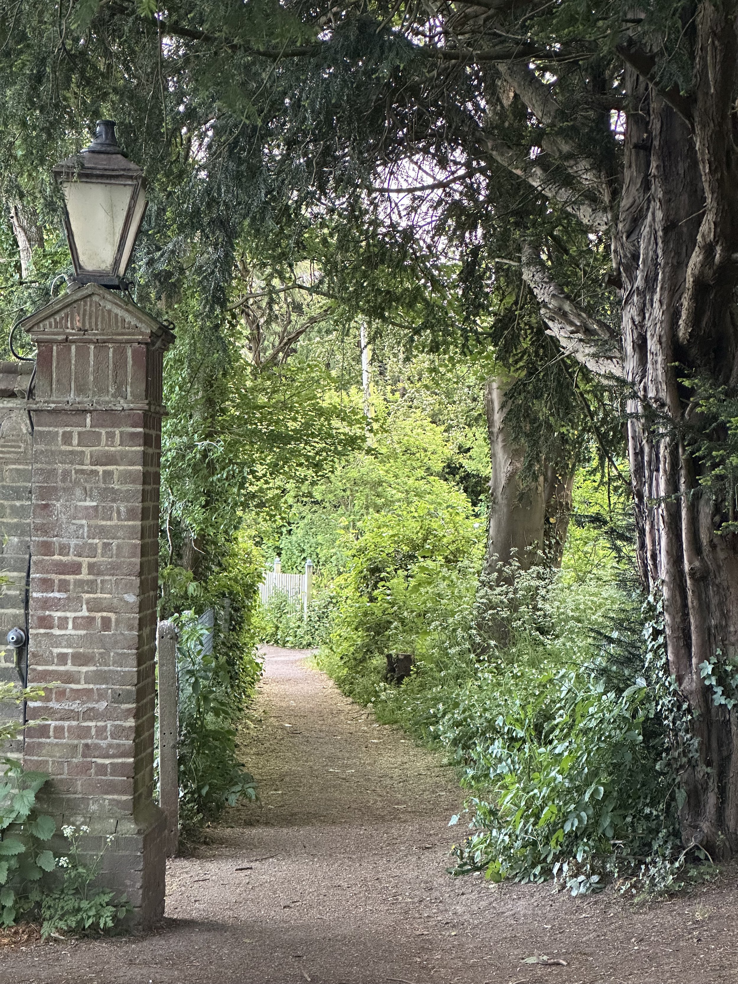 A garden pathway surrounded by lush green trees and bushes. A brick pillar with an old-fashioned lamp on top is on the left side of the pathway. The pathway is narrow and curves into the distance, lined with various plants and foliage.