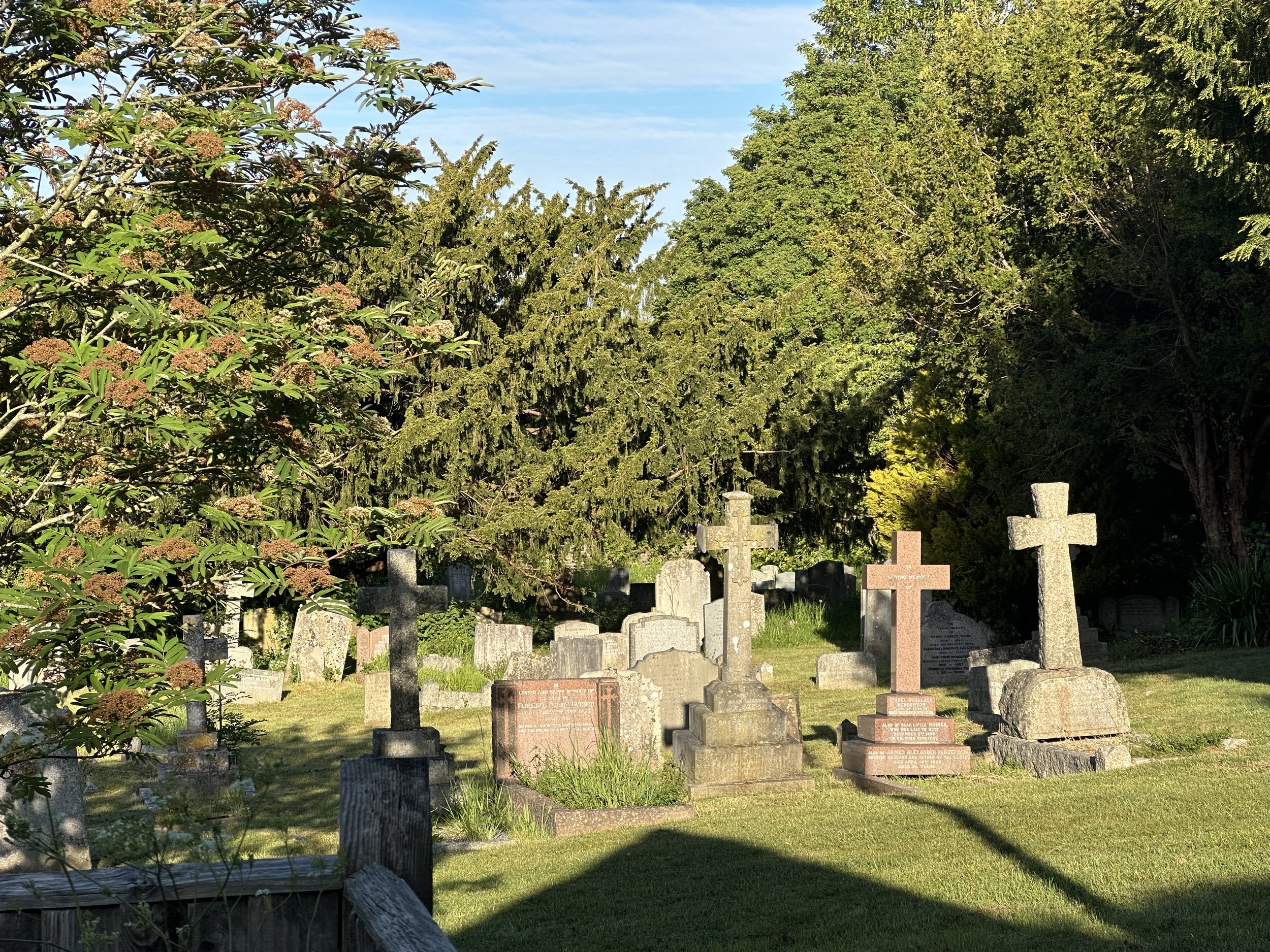 A graveyard with various tombstones and crosses surrounded by green trees and bushes under a partly cloudy blue sky.