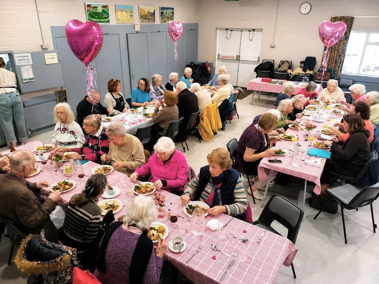 A gathering of elderly people enjoying a meal at multiple tables decorated with pink checkered tablecloths, pink heart-shaped balloons, and small flower arrangements in a community hall.