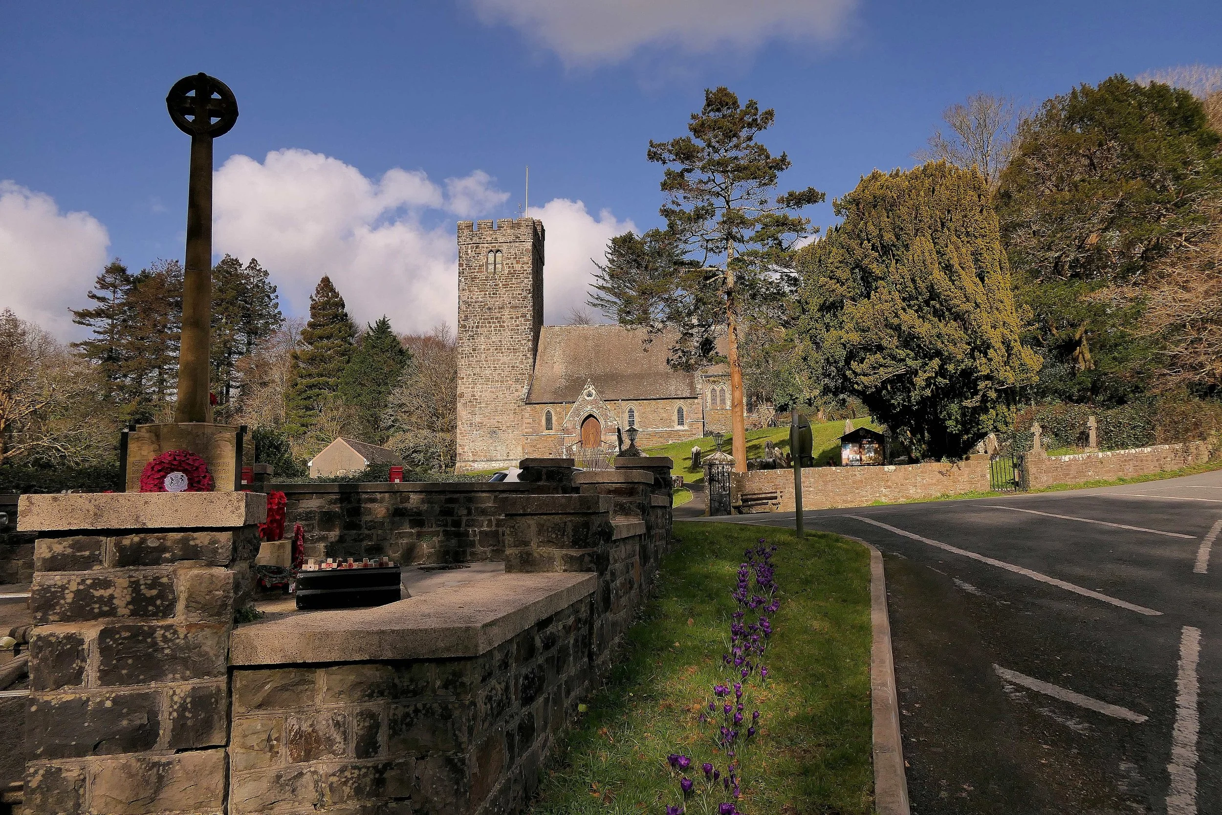A church with a stone tower surrounded by trees, a memorial with a cross, and a few flowers and benches in the foreground.
