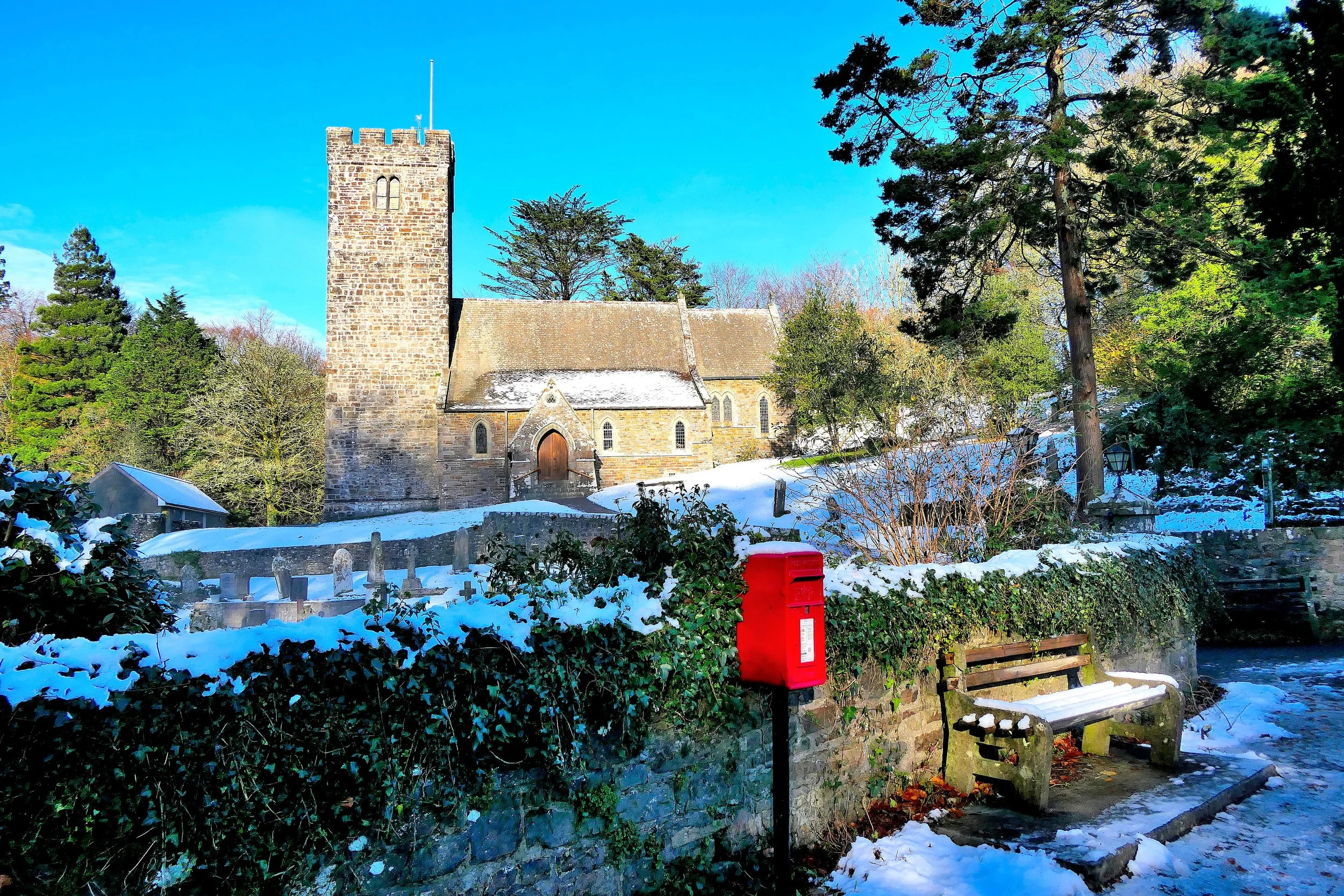 An old stone church with a tall tower surrounded by trees and snow on the ground, under a bright blue sky.