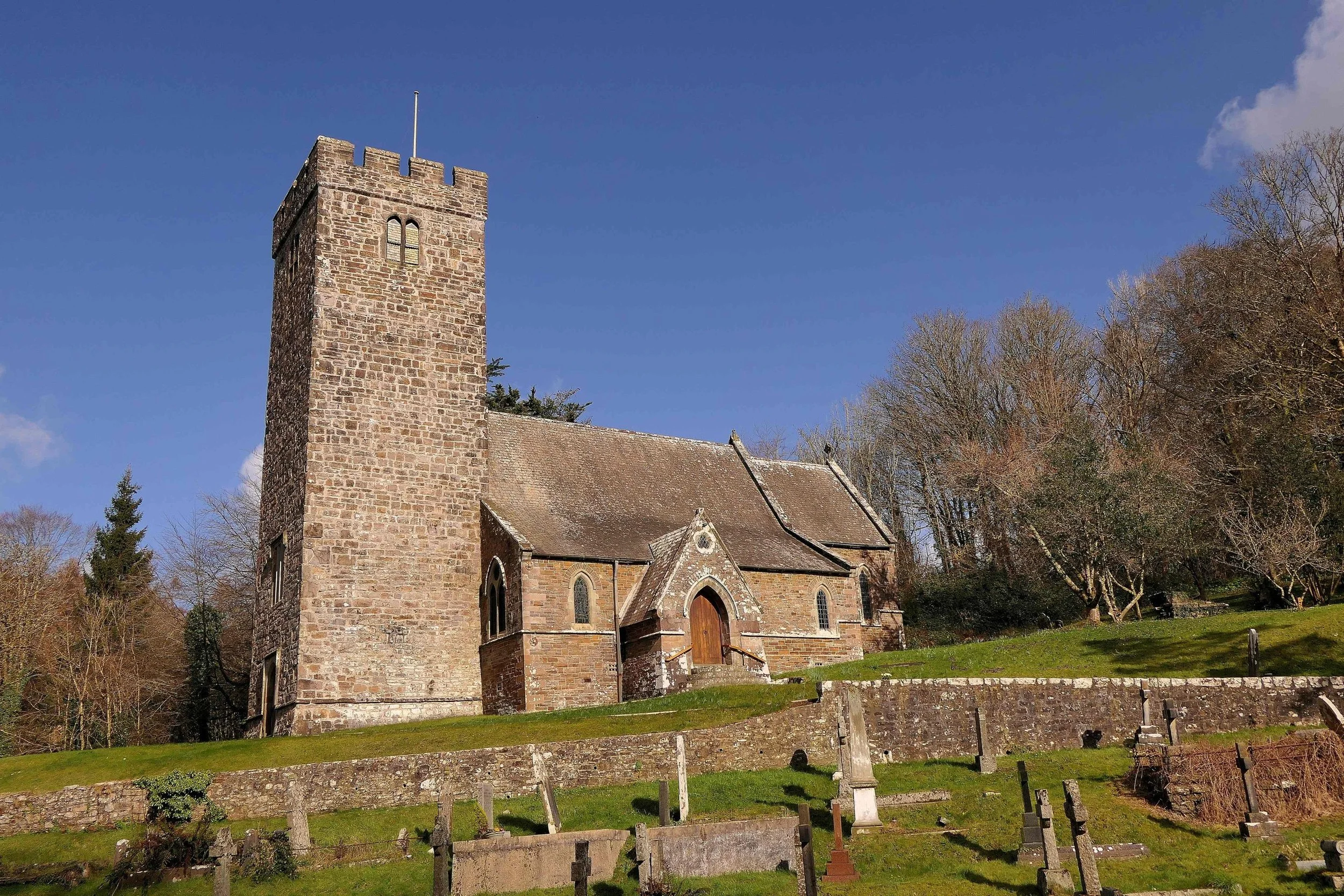 A historic stone church with a tall square tower, arched windows, a wooden door, and a graveyard in the foreground, surrounded by leafless trees under a blue sky.