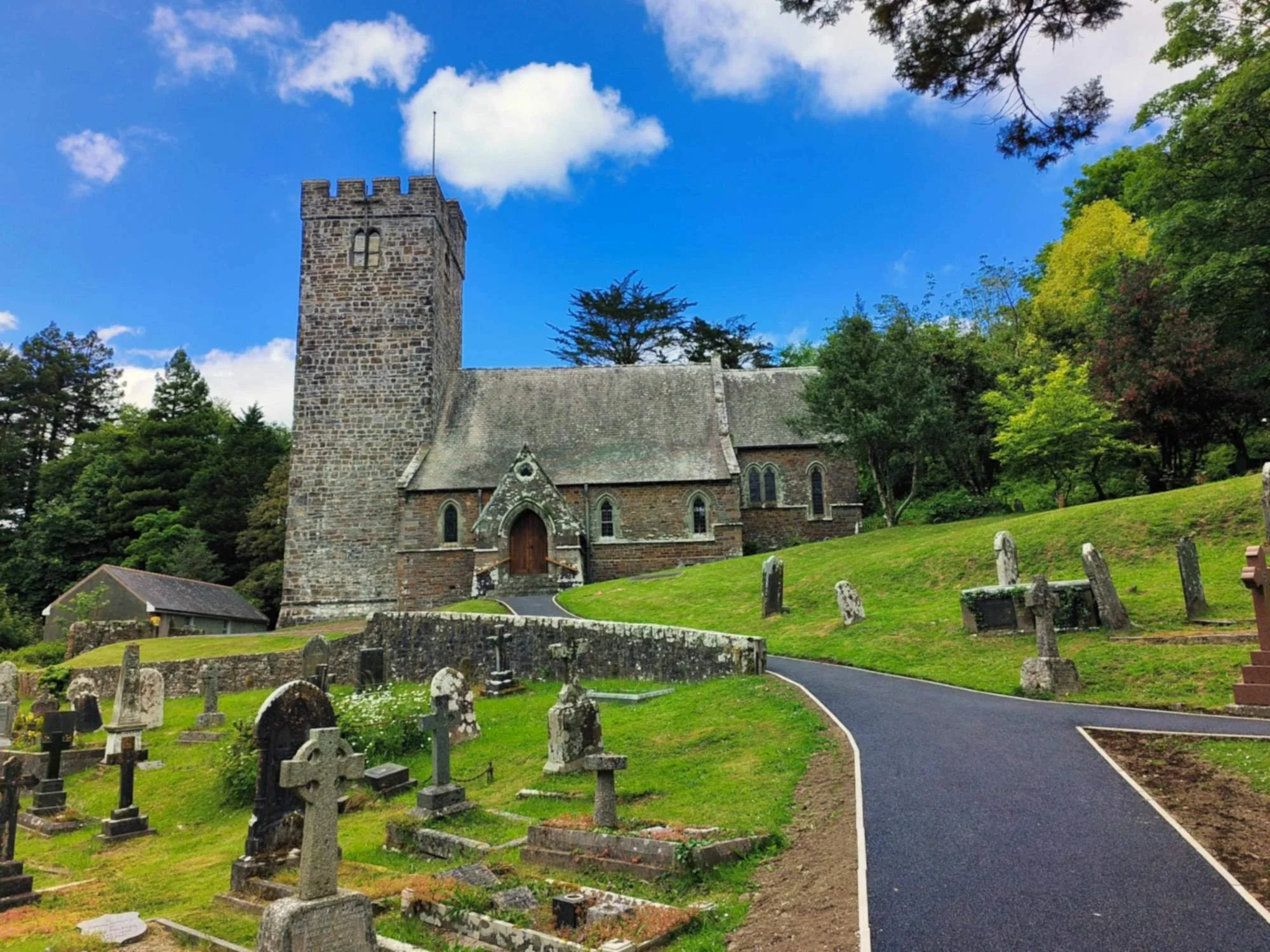 A stone church with a square tower and small arched windows, surrounded by a graveyard with old tombstones, on a lush green hill under a blue sky with scattered clouds and trees.