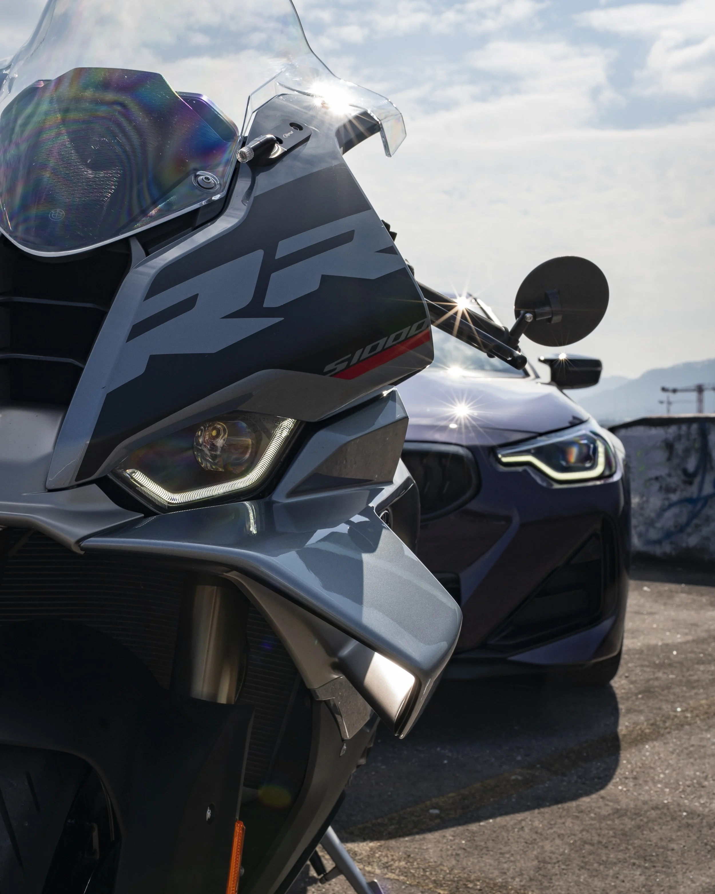Close-up of a grey and black motorcycle with a clear windshield, parked outdoors with other vehicles visible in the background under a partly cloudy sky.