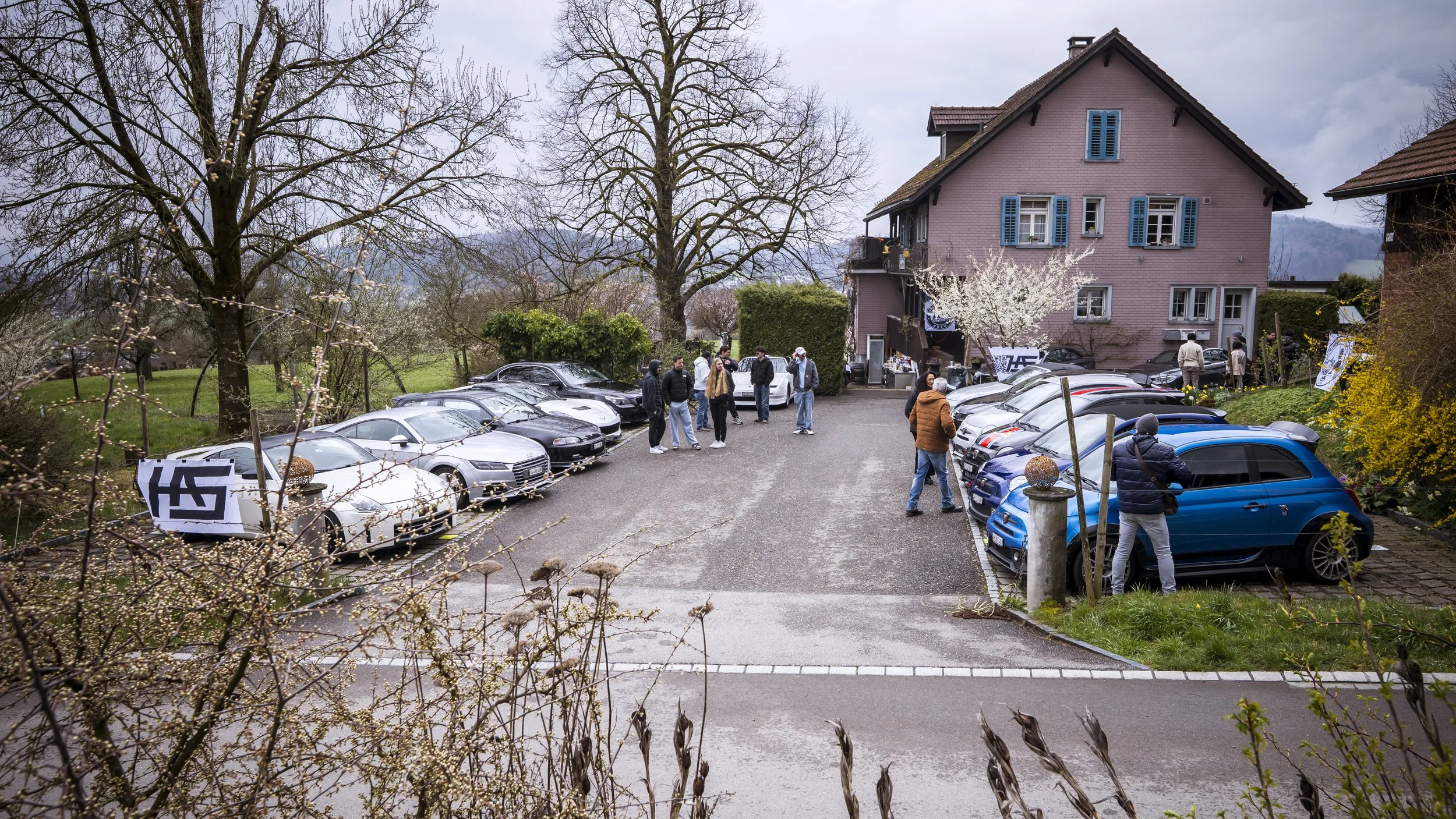 A parking lot in front of a house with people standing and talking. Several cars are parked, and there are trees with blossoming flowers around. The house is a pink, three-story building with blue shutters.