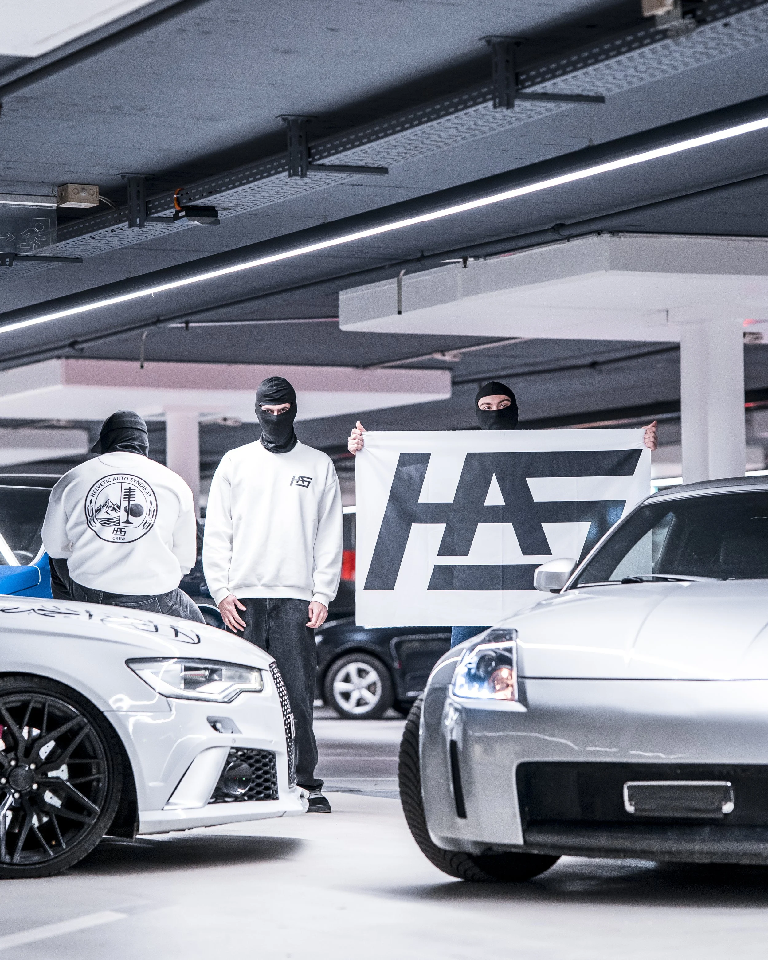 People dressed in black ski masks and white shirts holding a banner in a parking garage surrounded by parked cars.