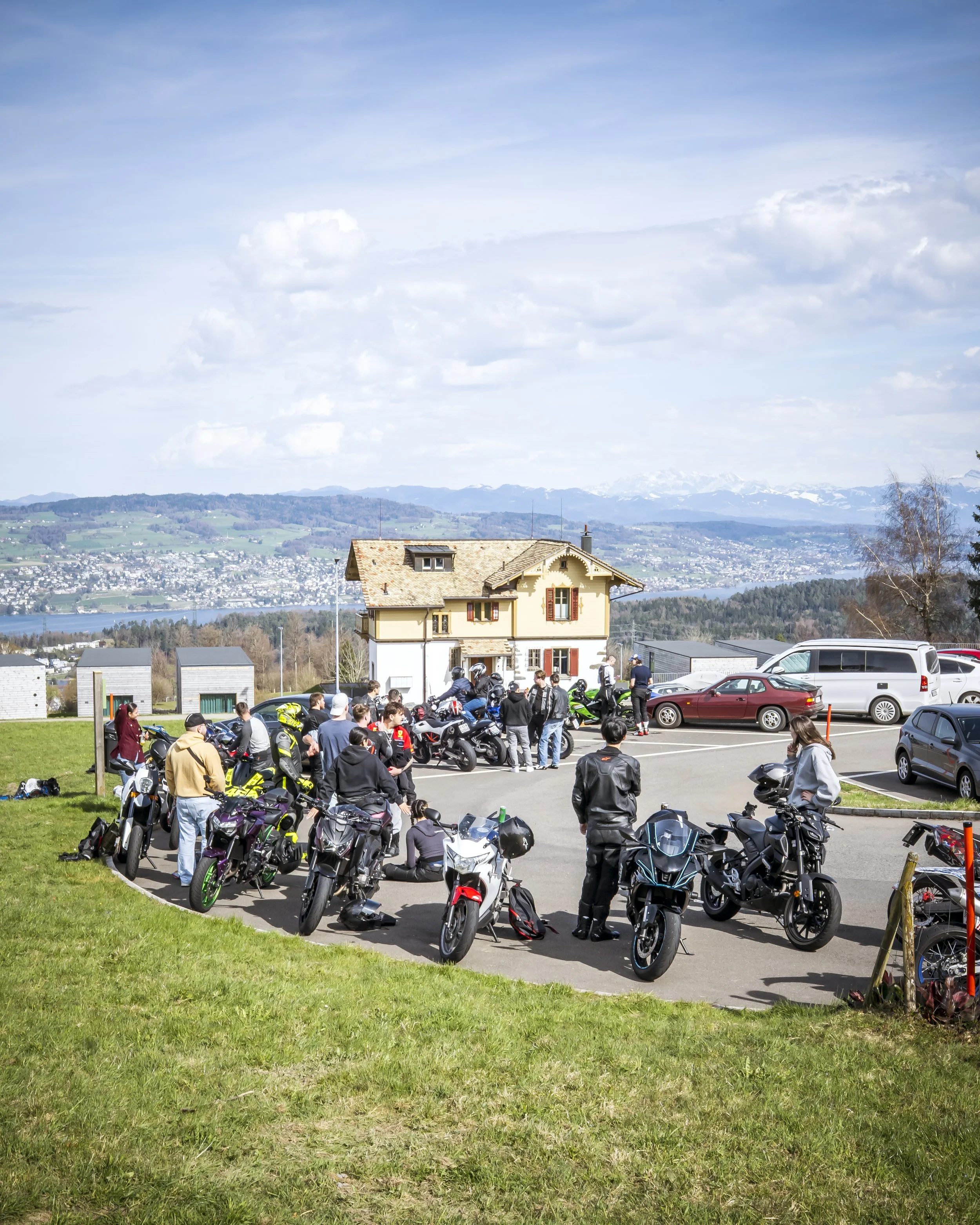 A parking lot with many motorcycles and a few cars, a group of people gathered around the motorcycles, a house on a hill in the background with a mountain range, partly cloudy sky, and a green field in the foreground.