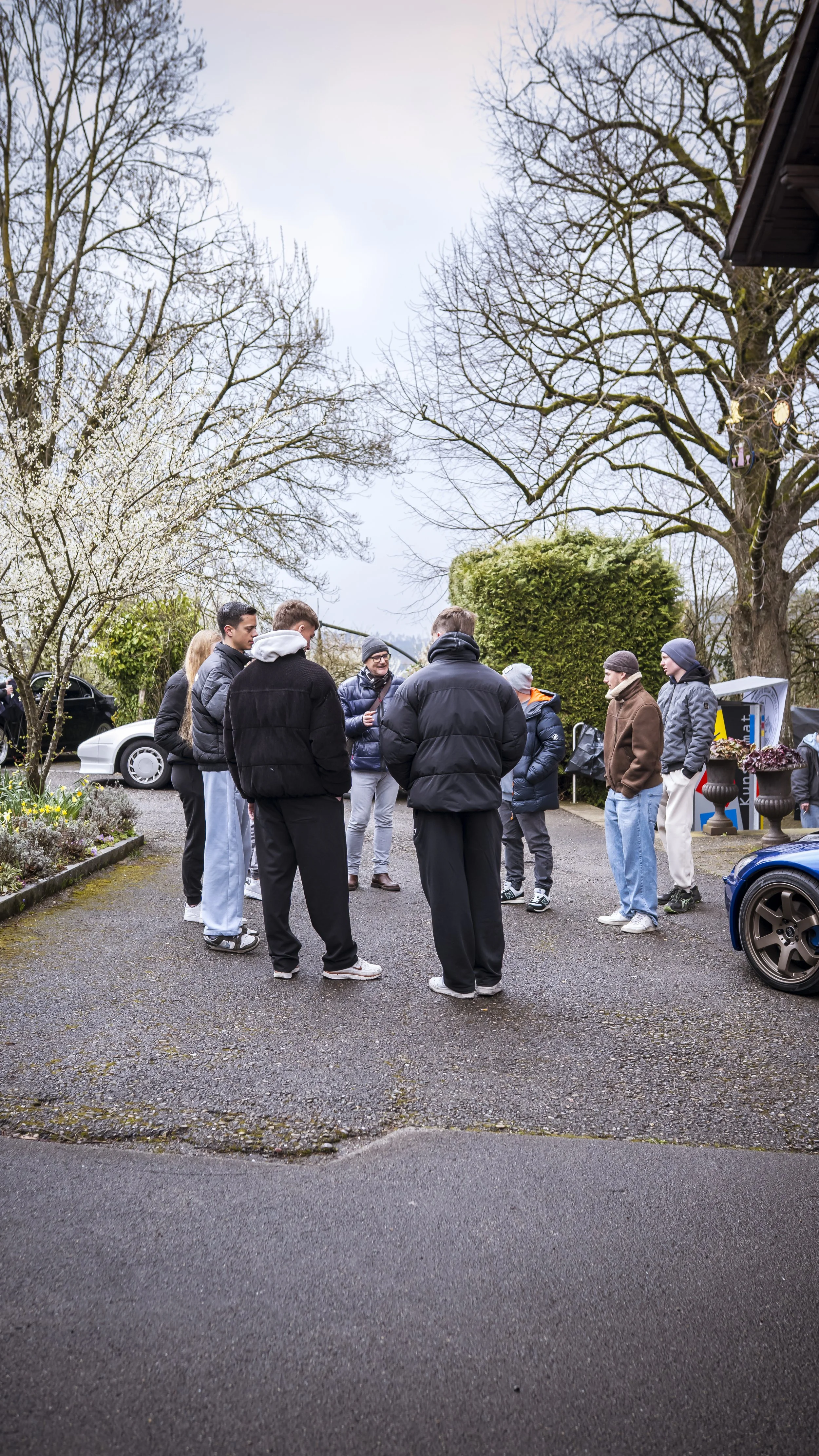 A group of young people standing outdoors on a cloudy day, engaged in conversation. They are wearing casual winter clothing and are gathered in a semi-circle. There are trees, some in bloom, and cars parked in the background.