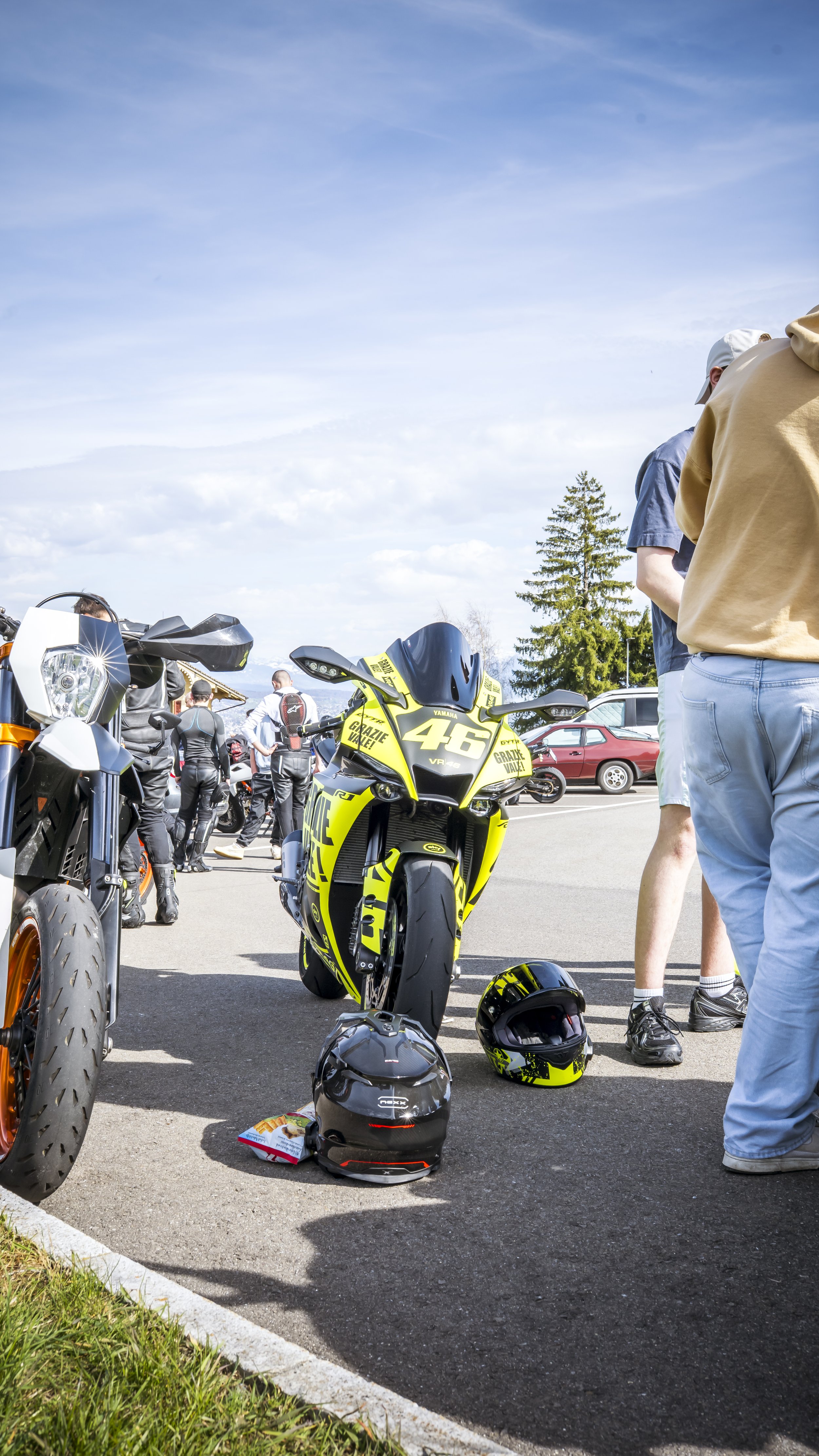 People gathered in a parking lot around motorcycles, helmets placed on the ground, with a clear sky and trees in the background.