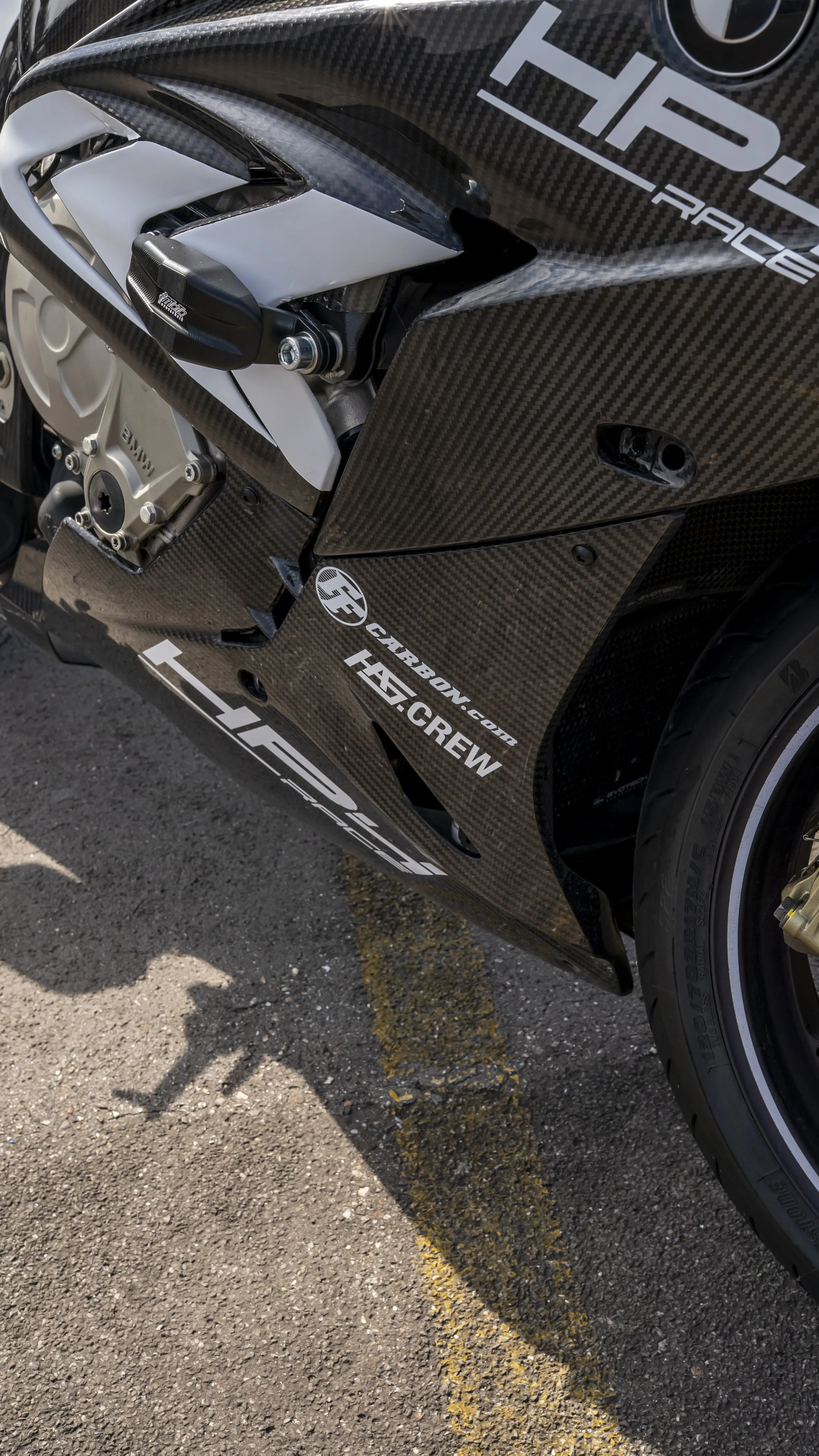Close-up of a racing motorcycle with black carbon fiber bodywork and silver components, parked on asphalt with yellow parking lines, featuring various sponsor stickers.