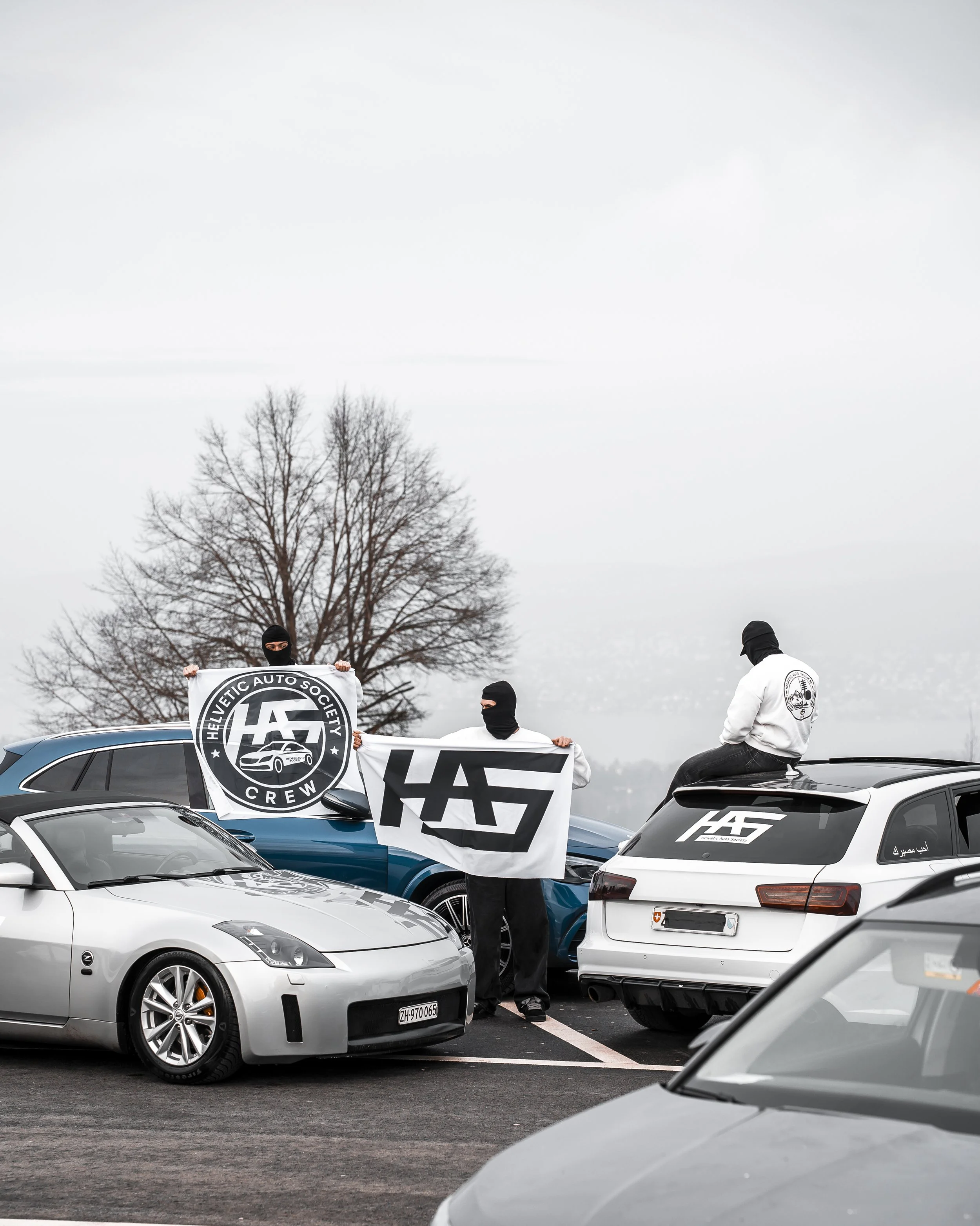 People wearing masks and holding banners with the Helvetic Auto Society logo, standing in a parking lot among parked cars, some cars with banners and logos, overcast sky, leafless trees in background.