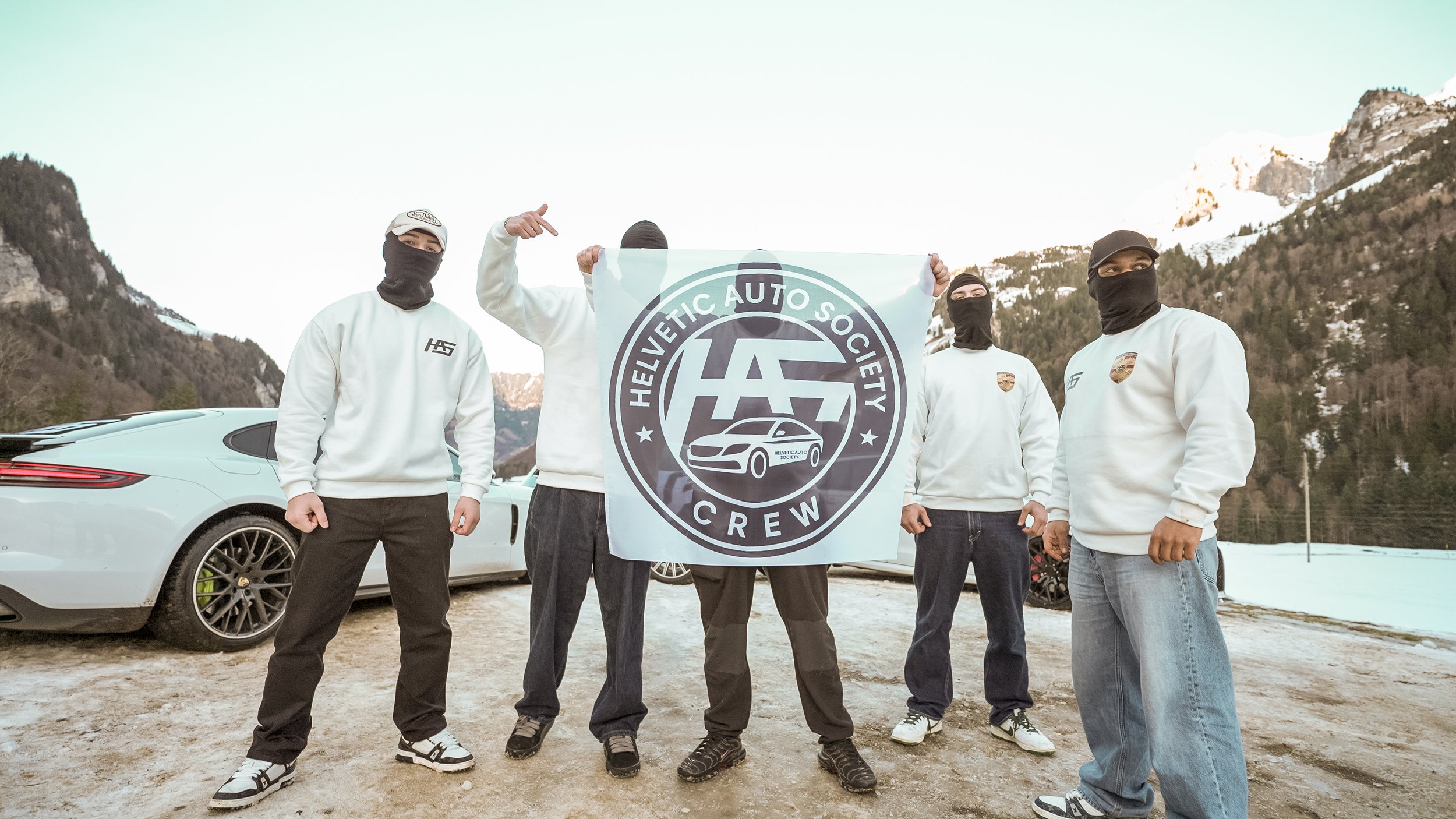 Group of five people wearing masks and white Helvetic Auto Society Crew sweatshirts, standing outdoors with mountains and snow in the background, holding a large flag with the society's logo and a car illustration.