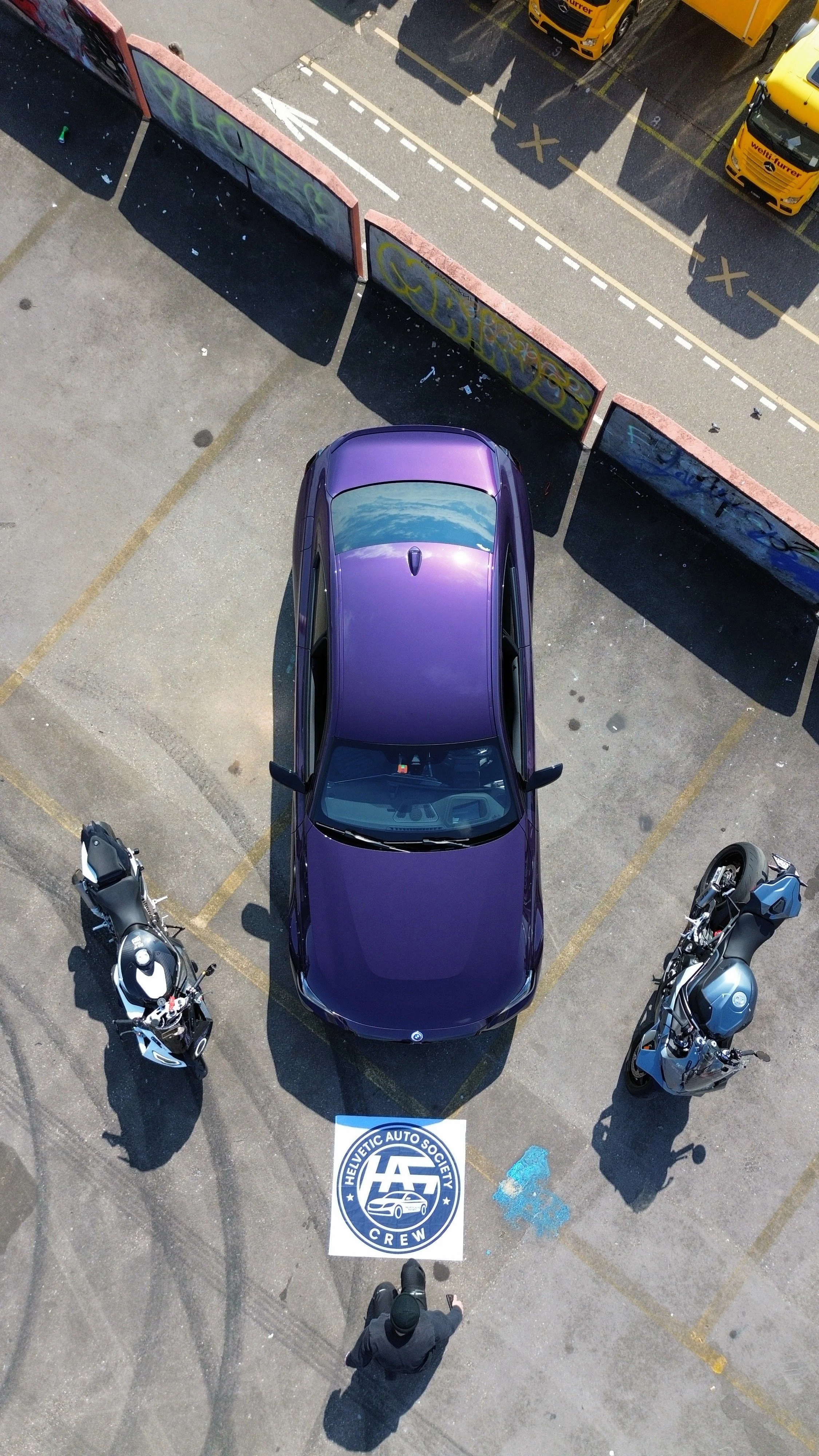 Overhead view of a parking lot with a purple car and two motorcycles, a man in dark clothing standing in front of a banner that reads 'HELICETIC AUTO SOCIETY CREW,' in front of graffiti-covered barriers and parked trucks.