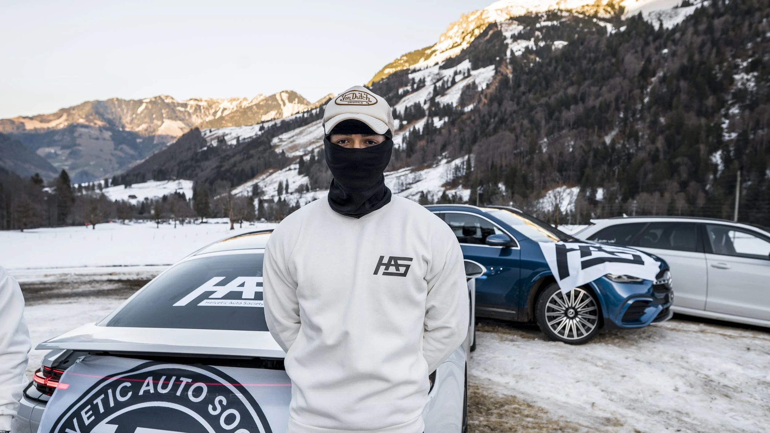 A person wearing a beige cap and black face covering standing outdoors in front of a mountain landscape and parked cars, including a white car with a partially visible logo reading  "HETIC AUTO SOCI".
