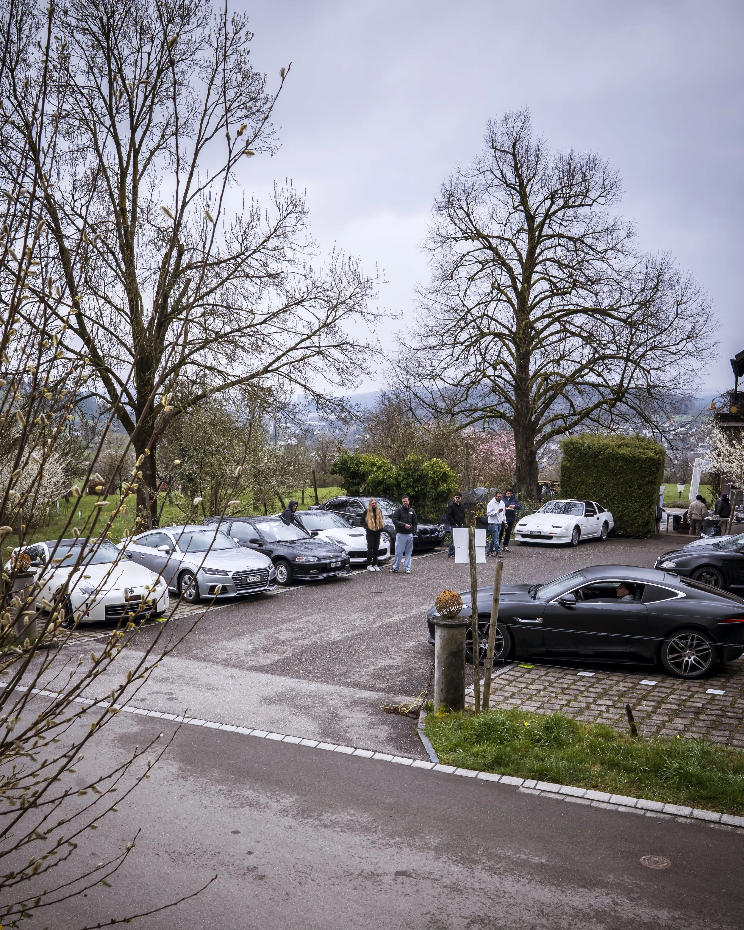 A parking lot filled with several luxury cars, including a white Lamborghini and a black BMW, with a group of people standing near the cars. In the background, there are large, leafless trees and a cloudy sky.