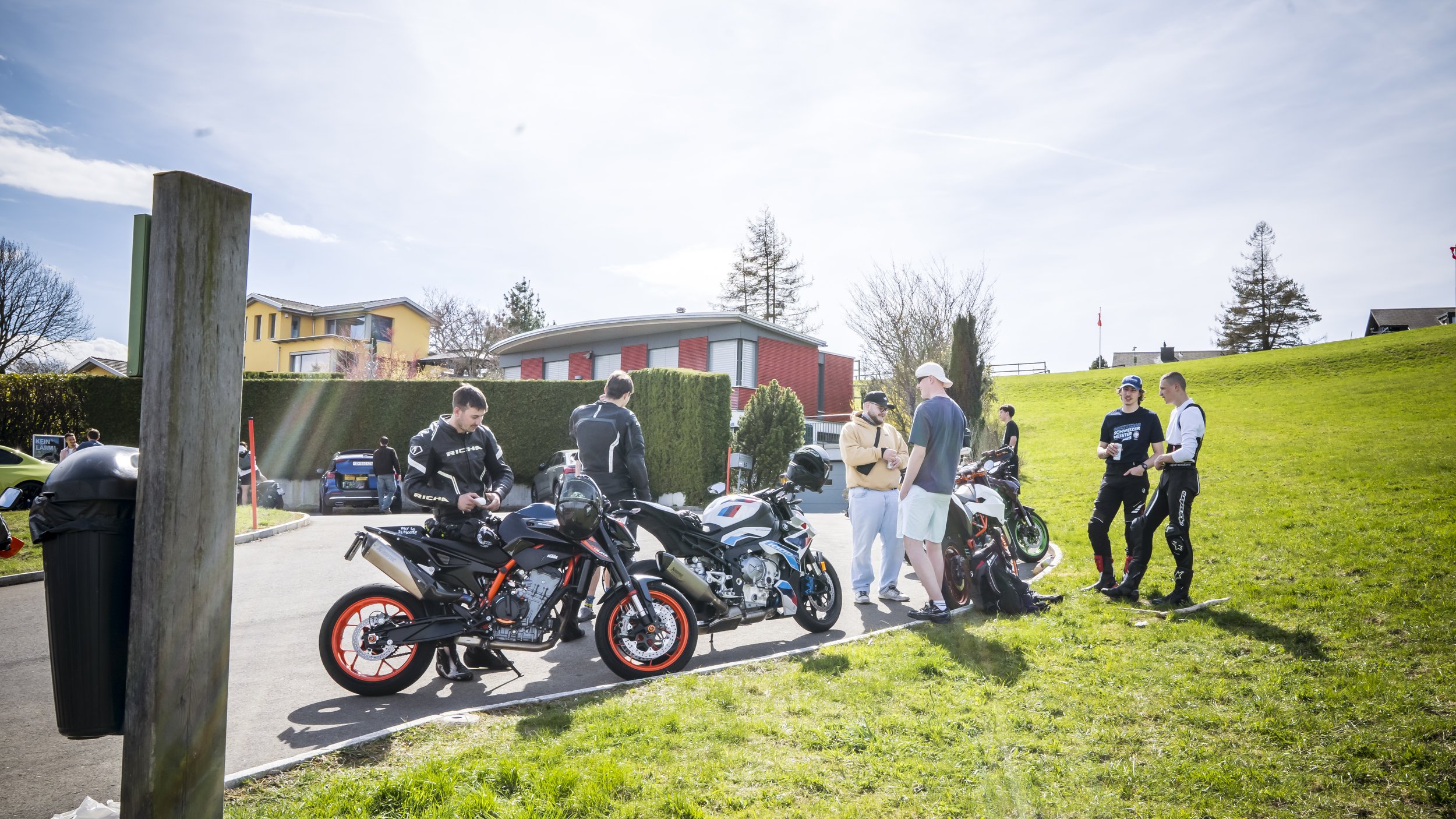 Group of people gathered outdoors with motorcycles, some wearing riding gear, in a grassy area near residential buildings on a sunny day.