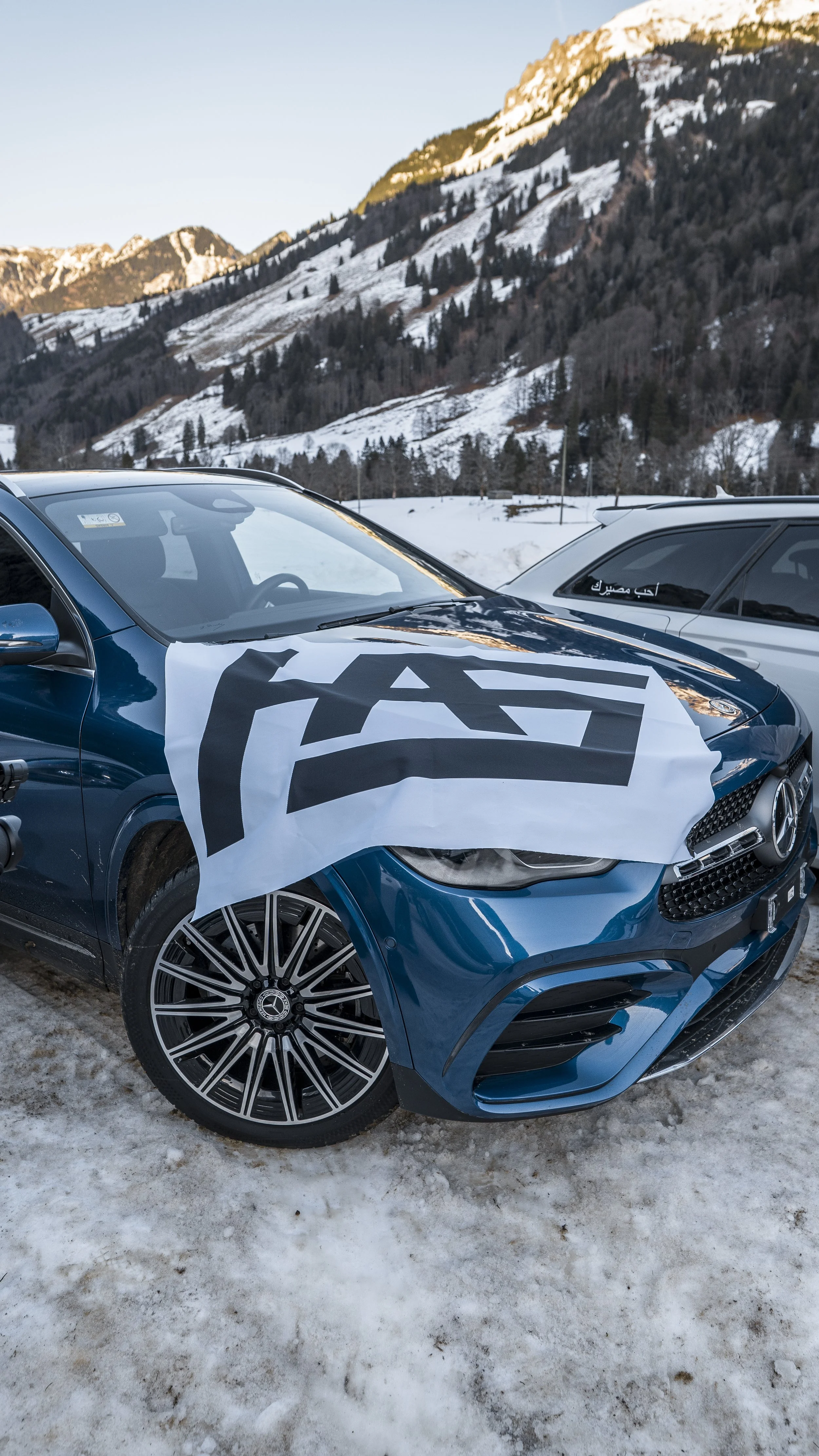 A blue Mercedes-Benz car with a large white flag on the hood displaying bold black text, parked on snowy ground with a mountainous landscape in the background.