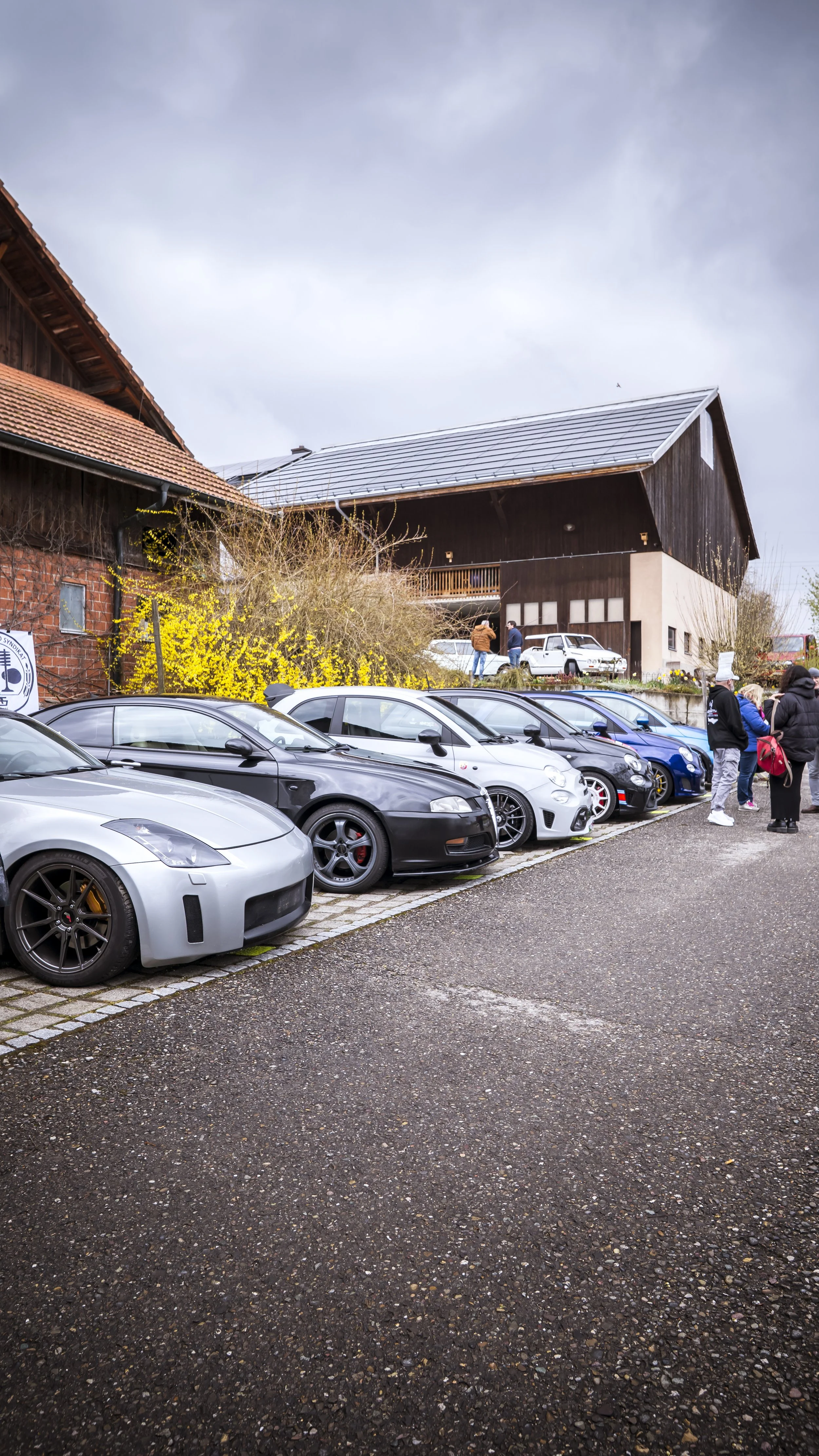 A parking lot with multiple sports cars parked in a row, a group of people standing and talking, a building with a sloped roof in the background, and cloudy sky above.