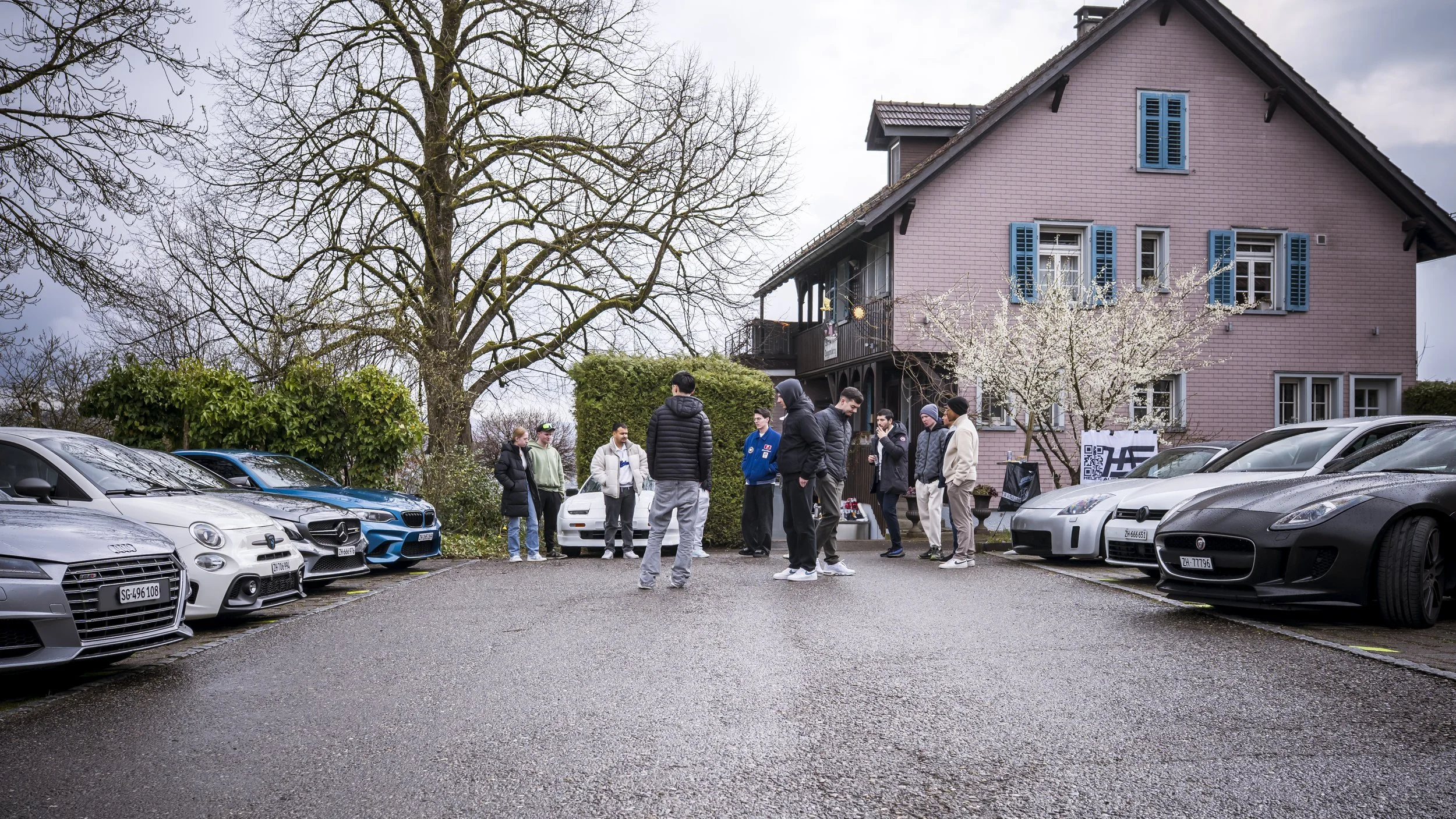 A group of people gathered outside a pink house with blue shutters, surrounded by parked cars, blooming trees, and leafless trees, on an overcast day.