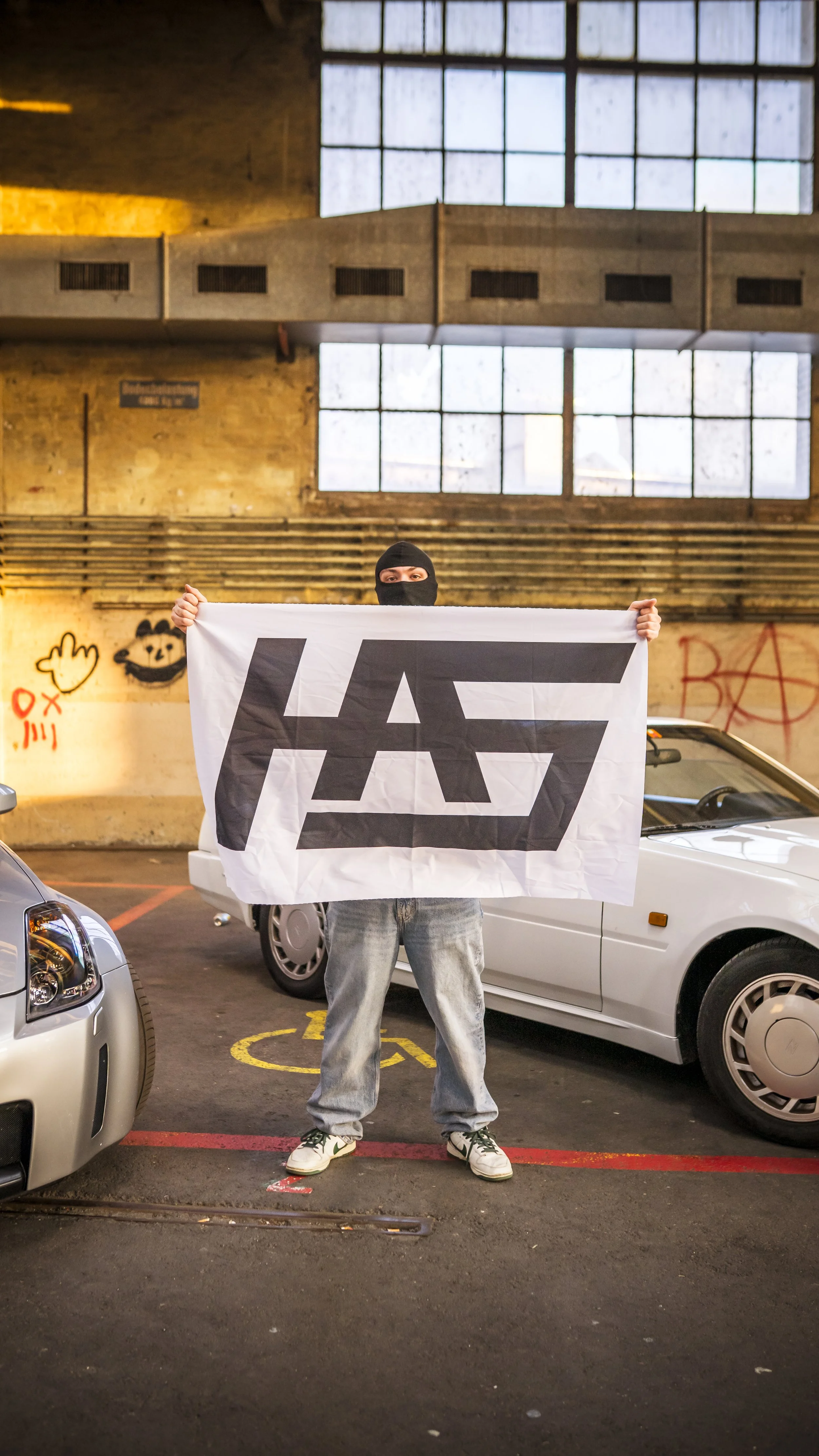 Person wearing a ski mask standing in a parking garage holding a flag with a stylized black logo on it.
