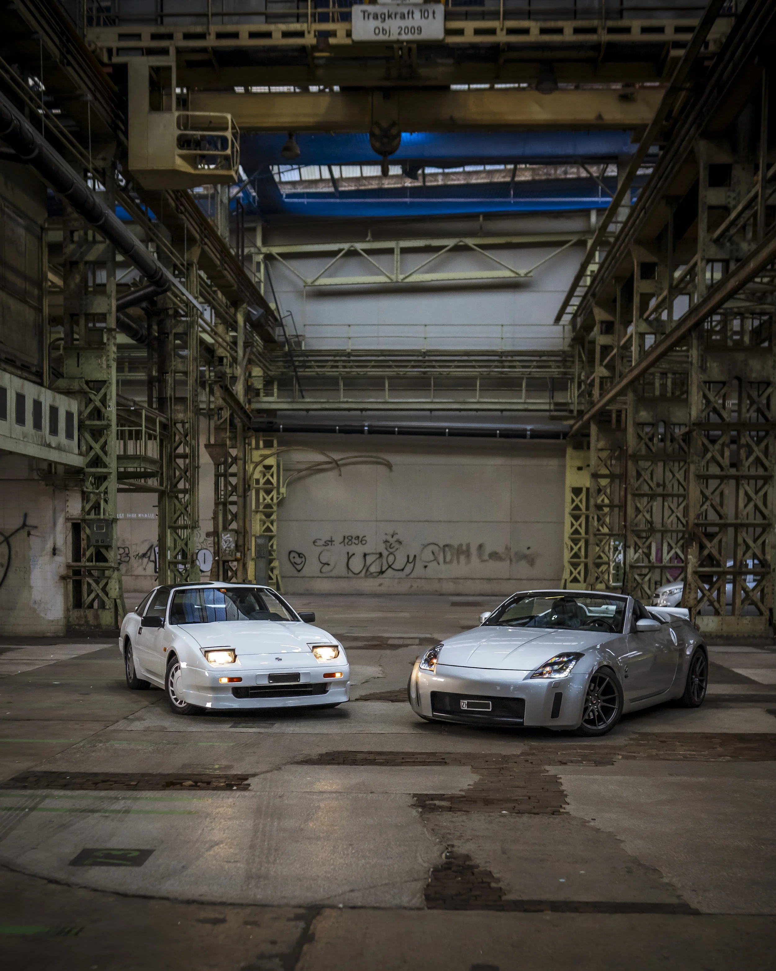 An industrial indoor parking area with three parked cars in the foreground, graffiti on the back wall, and a metal overhead structure with a sign that reads 'Tragkraft 10t OBJ 2009'.