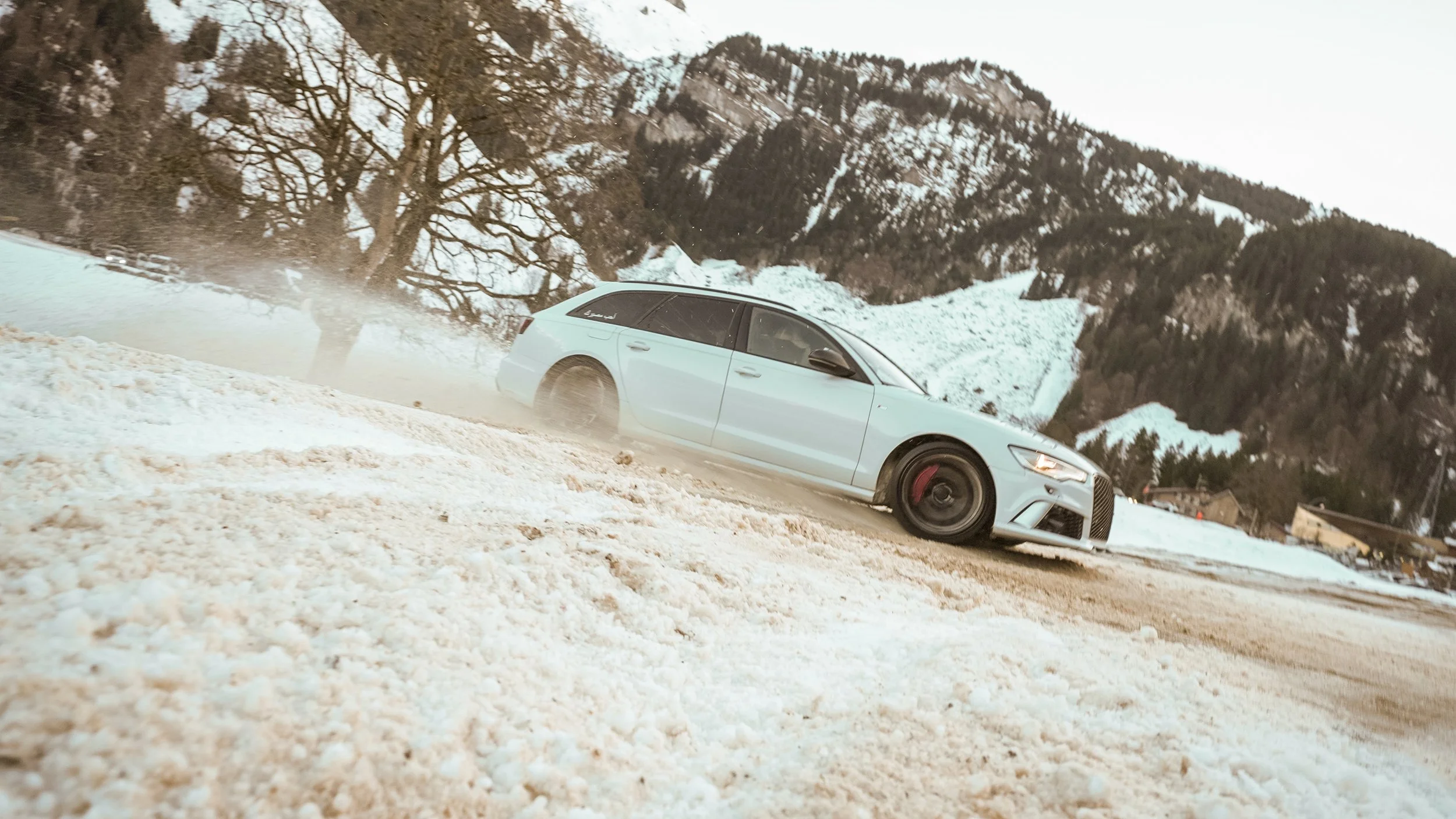 A white sports car driving on a snowy mountain road with snow-covered trees and mountains in the background.