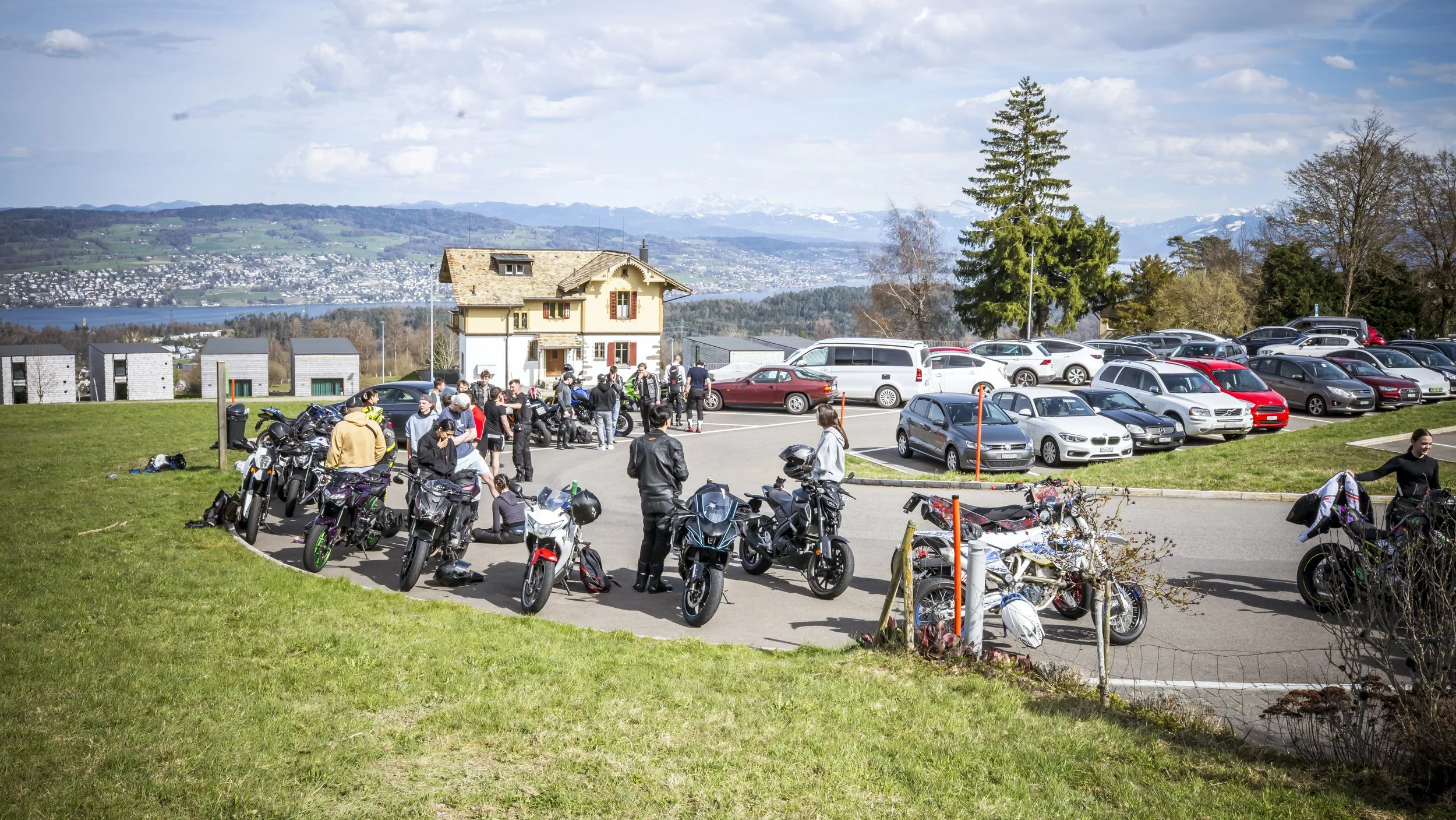 A parking lot filled with motorcycles and cars, with a group of people gathered near the motorcycles, a house in the background, and a scenic view of a lake and mountains in the distance.