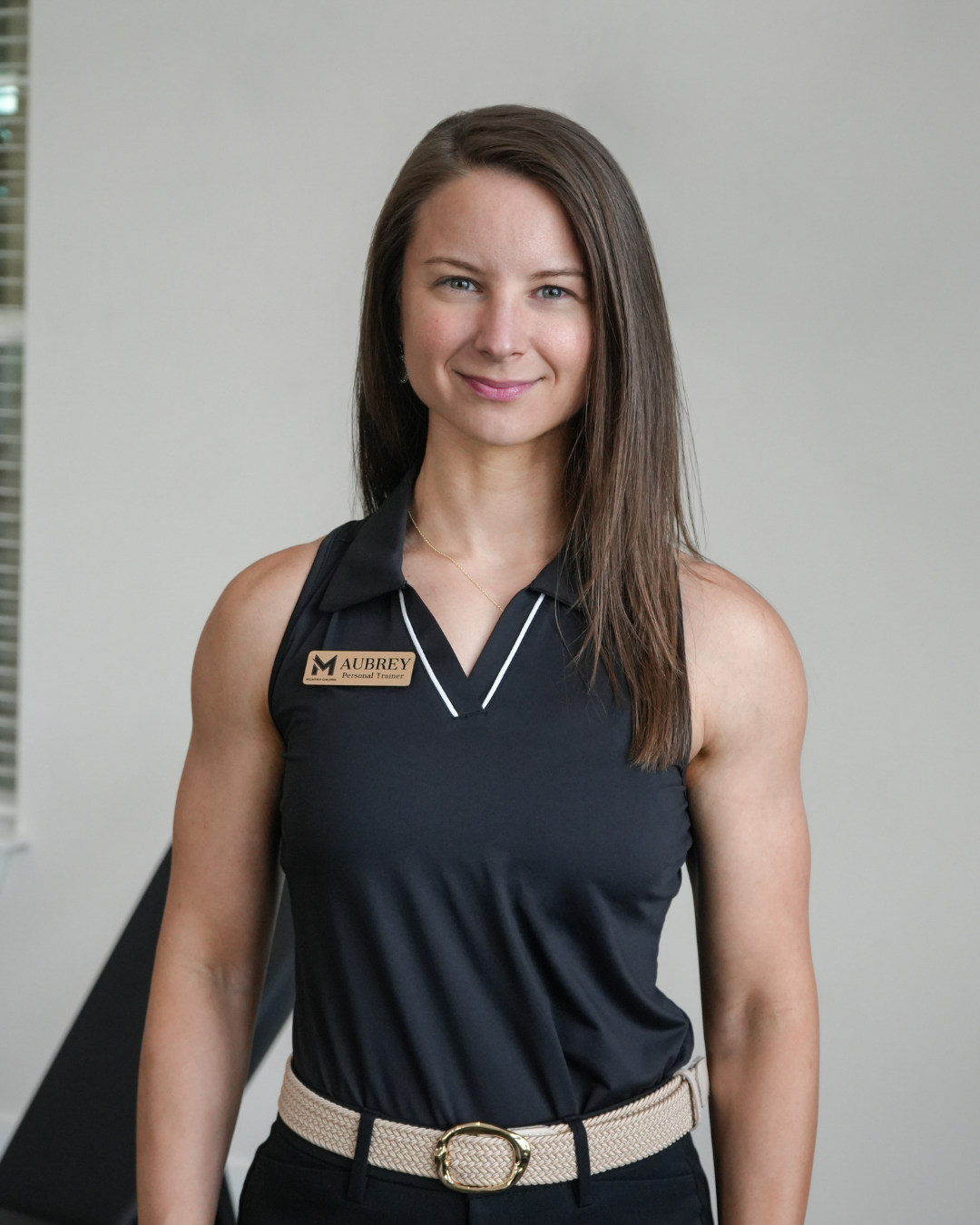 A woman with long brown hair wearing a black sleeveless polo shirt with a name tag that reads 'M Aubrey Personal Trainer,' standing indoors with a neutral background.