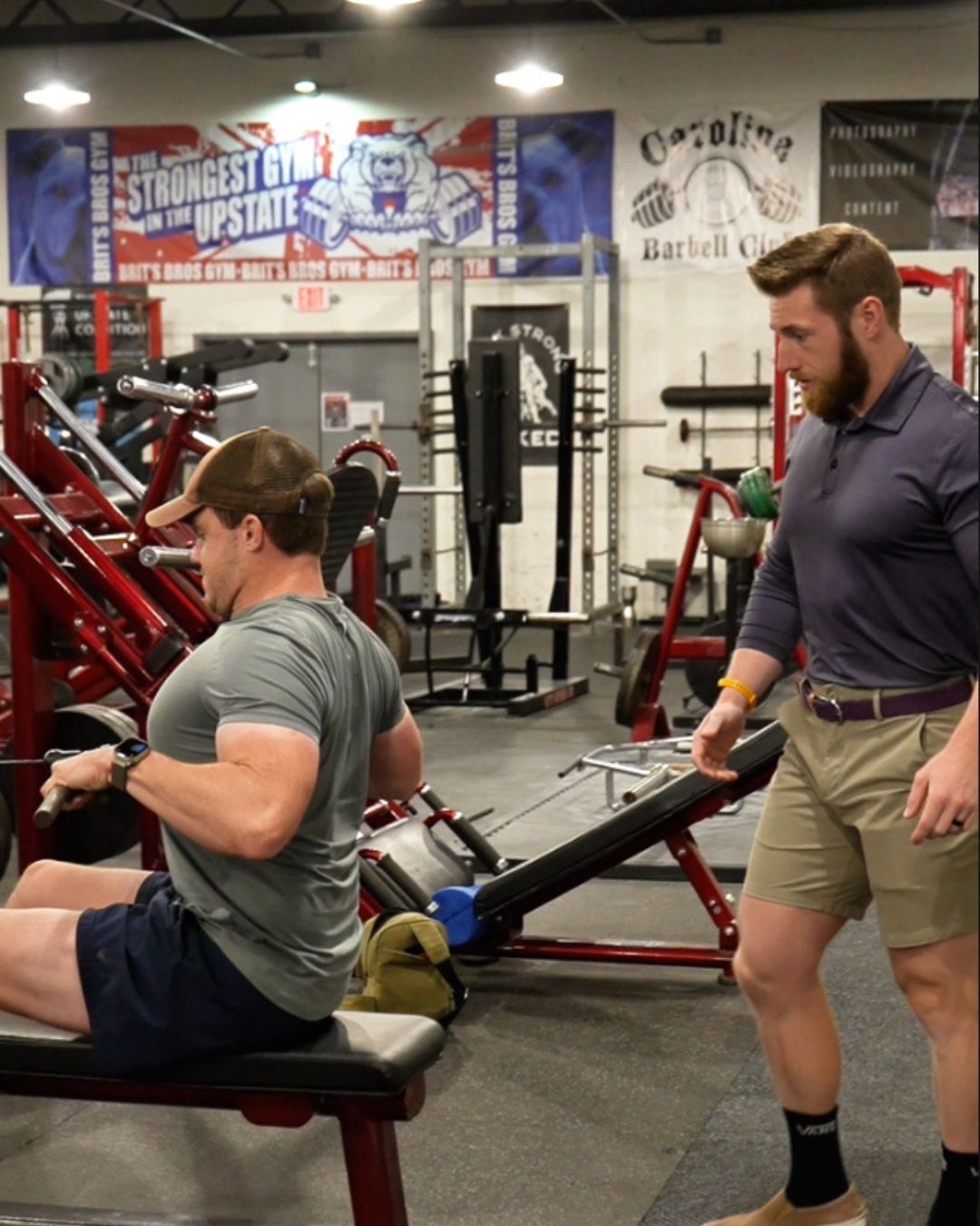 Two men in a gym, one sitting on a bench and the other standing nearby, surrounded by gym equipment and workout banners.