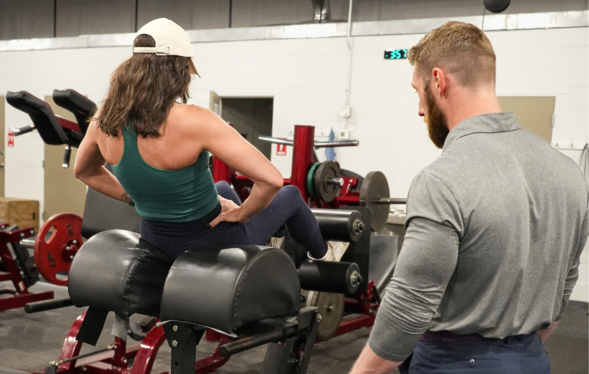 A woman wearing a white baseball cap, teal tank top, and dark leggings performs an ab exercise on a GHD machine while a professional male personal trainer in a gray shirt closely coaches her, observing her form.
