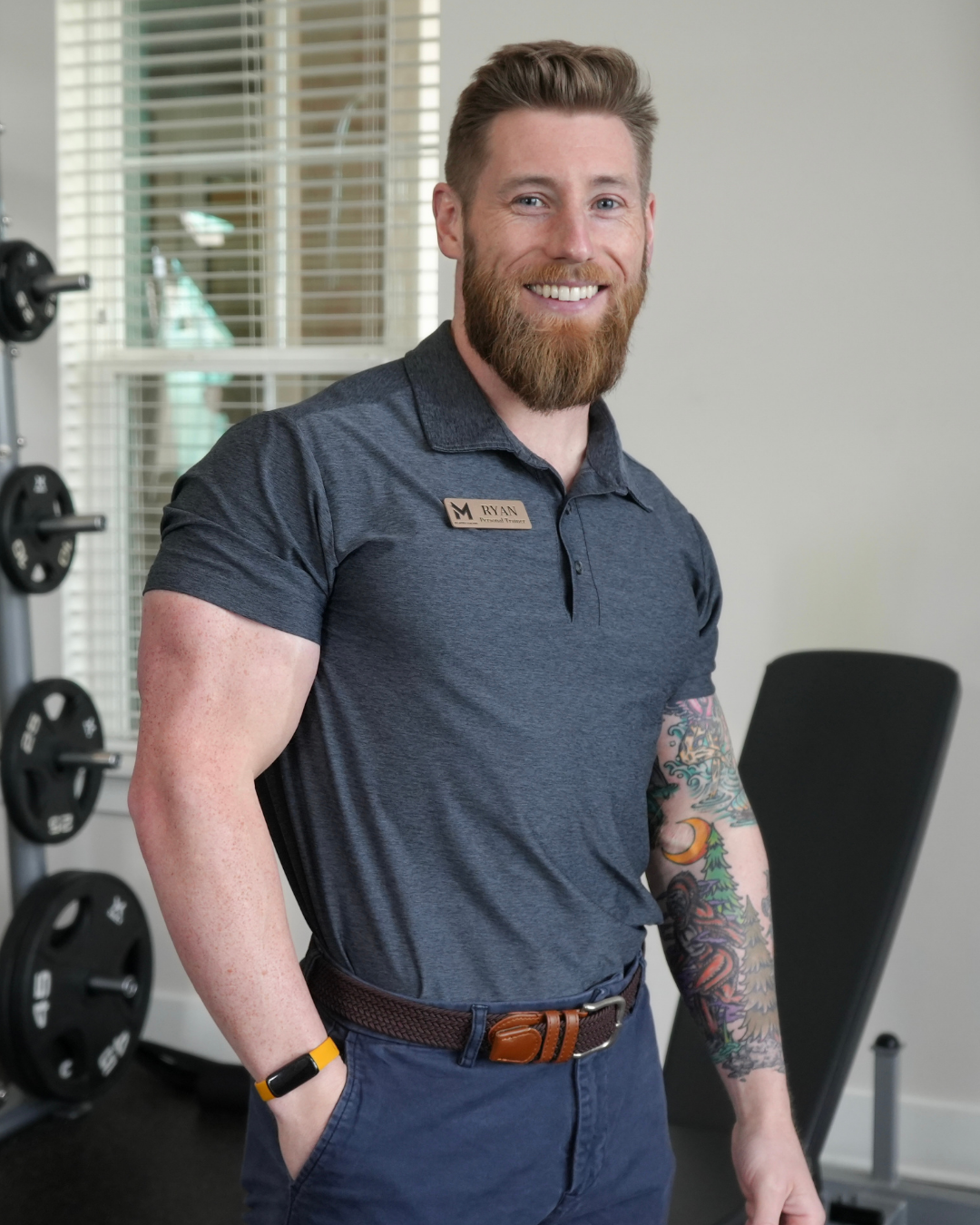 A smiling man with a beard, wearing a dark gray polo shirt with a name tag that reads "Ryan," and an arm tattoo, standing in a gym or fitness studio.