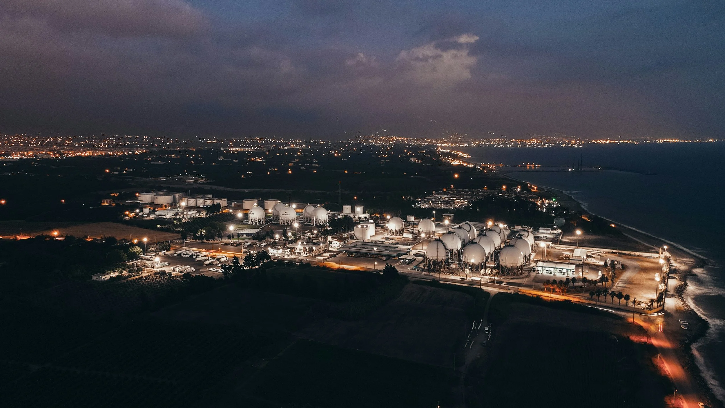 Aerial view of a coastal industrial area at night with illuminated storage tanks and bright city lights in the distance under a cloudy sky.