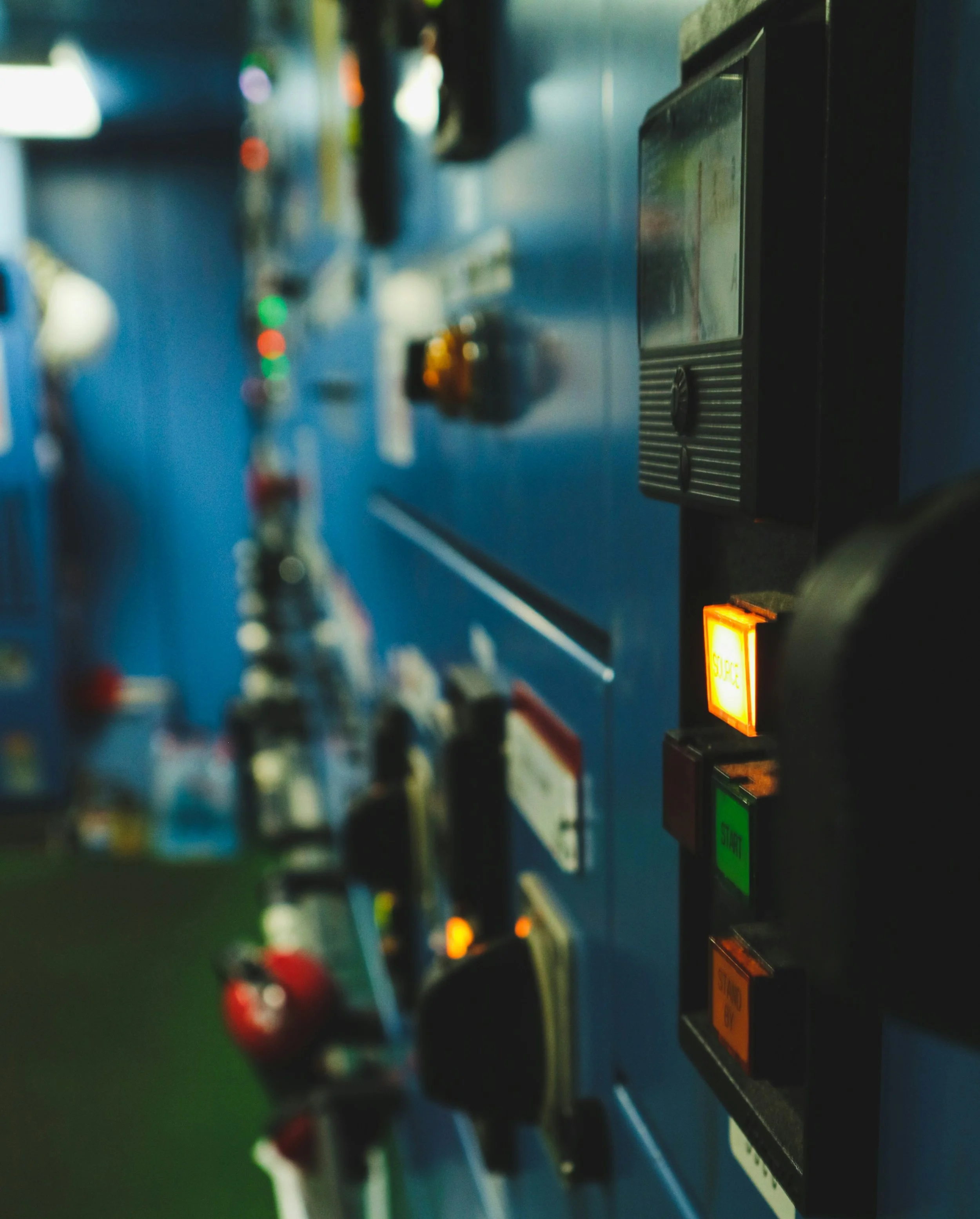 Close-up of a control panel with colorful switch buttons and indicator lights mounted on a blue surface, with vague background elements.