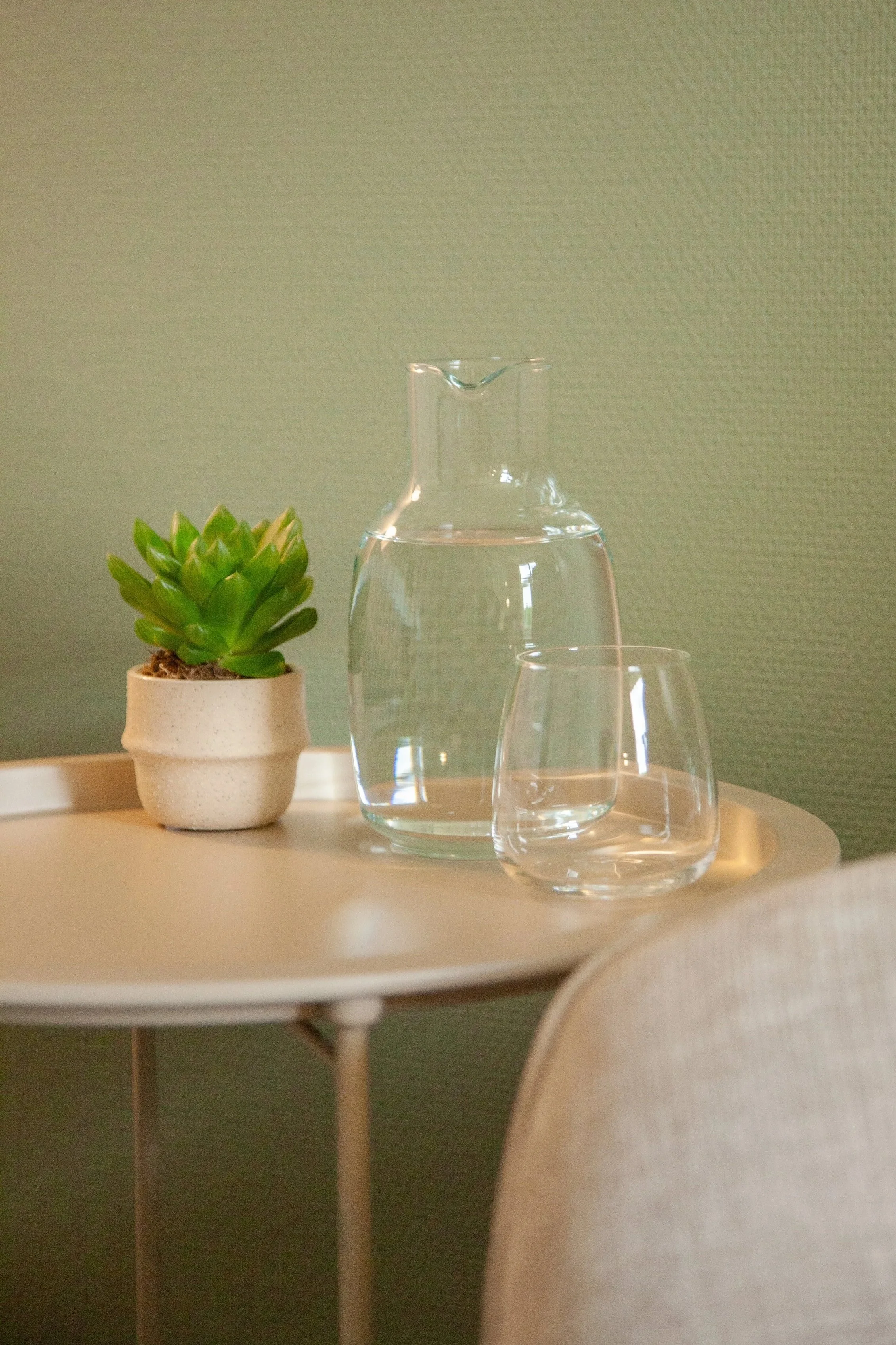 En liten grön växt i en beige kruka framför ett klart glas och en vattenkaraff på en ljus bordskant med en grön vägg i bakgrunden.