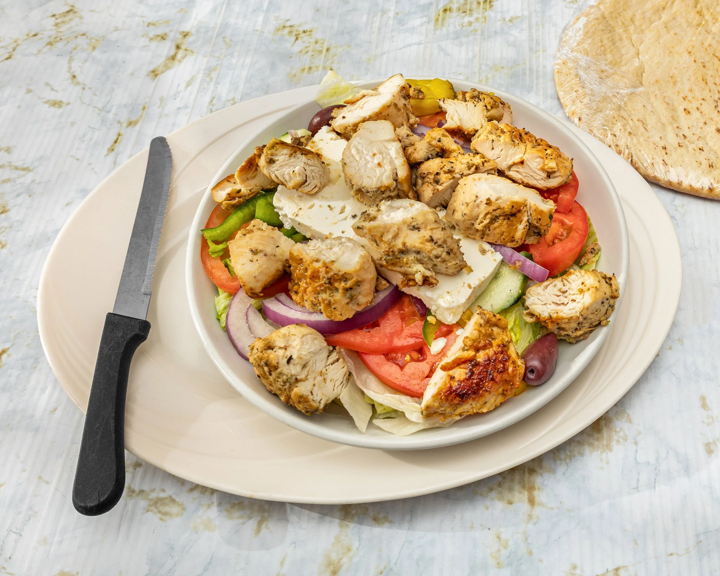 A plate with grilled chicken pieces, sliced tomatoes, lettuce, cucumber, purple onions, and feta cheese. There is a sandwich loaf wrapped in plastic on the side, and a serrated knife on the plate.