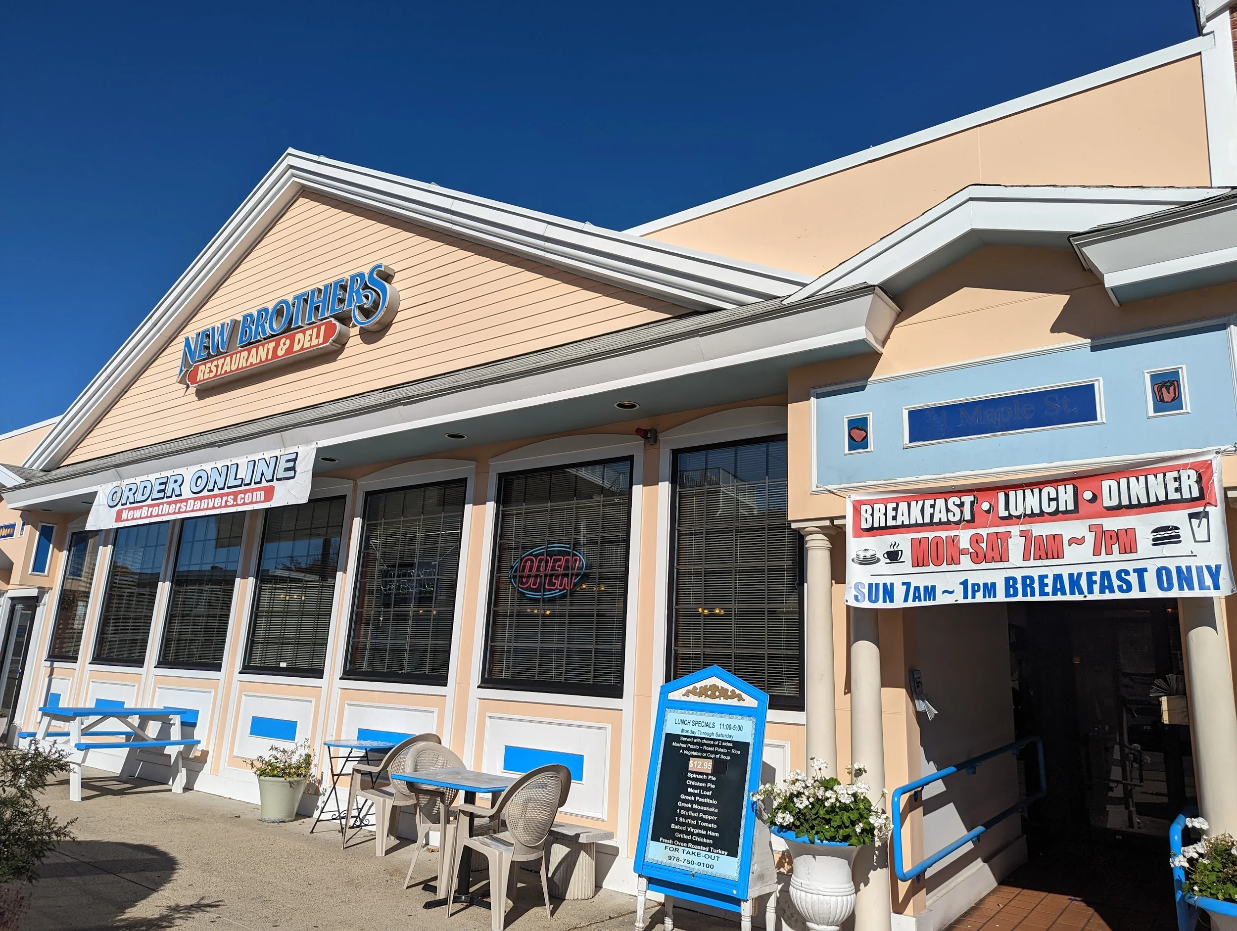 Exterior of a restaurant named New Brothers Restaurant & Deli with outdoor seating, umbrellas, and signage displaying menu and hours, under a clear blue sky.