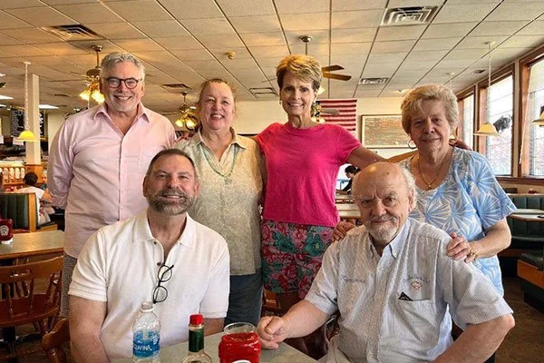 Group of six smiling people, three men and three women, gathered in a restaurant with wooden tables and chairs, American flag in the background, enjoying a meal.
