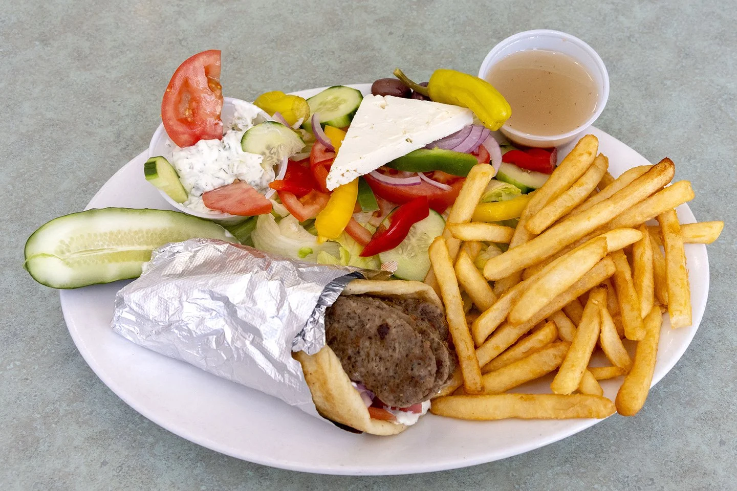 Plate with French fries, Greek salad, pita with lamb kebab, cucumber slices, tomato, onion, feta cheese, pepperoncini, and dressing.