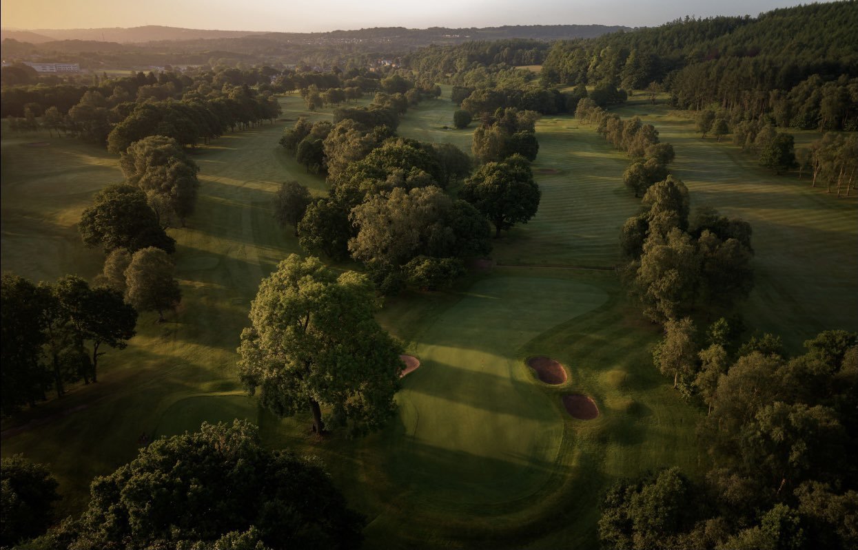 Aerial view of a golf course during sunset, with green fairways, sand bunkers, and scattered trees.